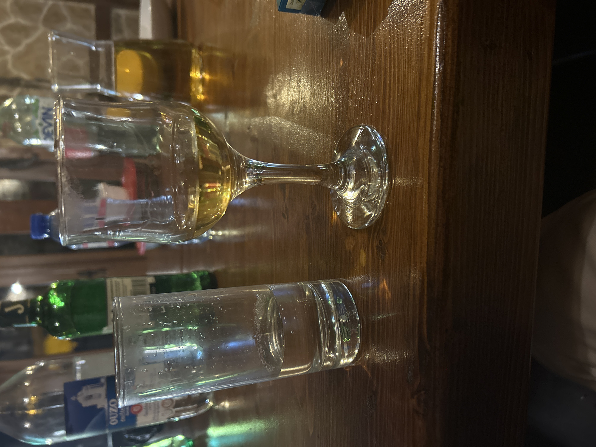 The image shows a close-up of a wooden table with several glasses and bottles on it. One glass contains a yellow liquid, possibly wine or a similar beverage. Other glasses are empty. The bottles appear to contain various drinks. The image was taken in Mirovyane, Bulgaria.