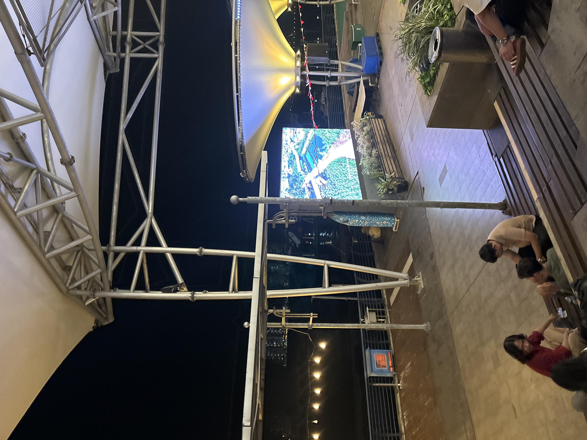 The image depicts an outdoor seating area at night, likely a public space or entertainment venue, possibly located along the waterfront in North Jakarta, Indonesia. The scene is dominated by a large, white fabric canopy supported by metal scaffolding. In the background, a bright LED screen displays an aerial view of a city with waterways and greenery.
Several people are visible in the foreground and middle ground. Three individuals are seated on wooden steps, appearing to be engaged in conversation or a casual activity. One person, wearing a maroon top and tan pants, is seated on the left, reaching towards a small object on a bench. Another person, in a light brown shirt and dark pants, is seated to their right, looking down. A third individual, in a green shirt, is partially visible between them, also seated. Further back, near the LED screen, several planters filled with greenery are arranged.
The environment appears to be an elevated walkway or plaza with wooden decking and railings. String lights are visible in the distance, illuminating the dark water. A blue bin and a trash receptacle are also present. The overall impression is one of a relaxed evening gathering in an urban setting.