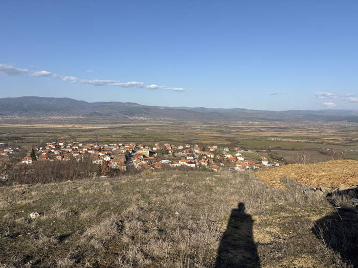 The image presents a panoramic, high-angle view overlooking the village of Novo Leski, Bulgaria, and its surrounding environment under clear daylight. The foreground consists of dry, unkempt grass and sparse, brown vegetation covering an uneven hillside. A distinct, elongated human shadow, cast by the photographer, extends across the lower-right quadrant of this foreground.
The midground is dominated by the village itself, which comprises numerous residential buildings, largely characterized by orange-red tiled roofs. These structures are densely clustered, interspersed with dormant trees and shrubs. Beyond the village, a wide, flat valley stretches into the distance, appearing as a patchwork of brown and green agricultural fields and undeveloped land.
The background is defined by a range of moderate-sized mountains with visible tree cover, forming the distant horizon. The sky above is bright, clear blue with a few wispy white clouds scattered across the upper portions. The scene is illuminated by direct sunlight, indicated by the prominent shadow in the foreground. No other individuals or specific actions are discernible within the visible landscape.