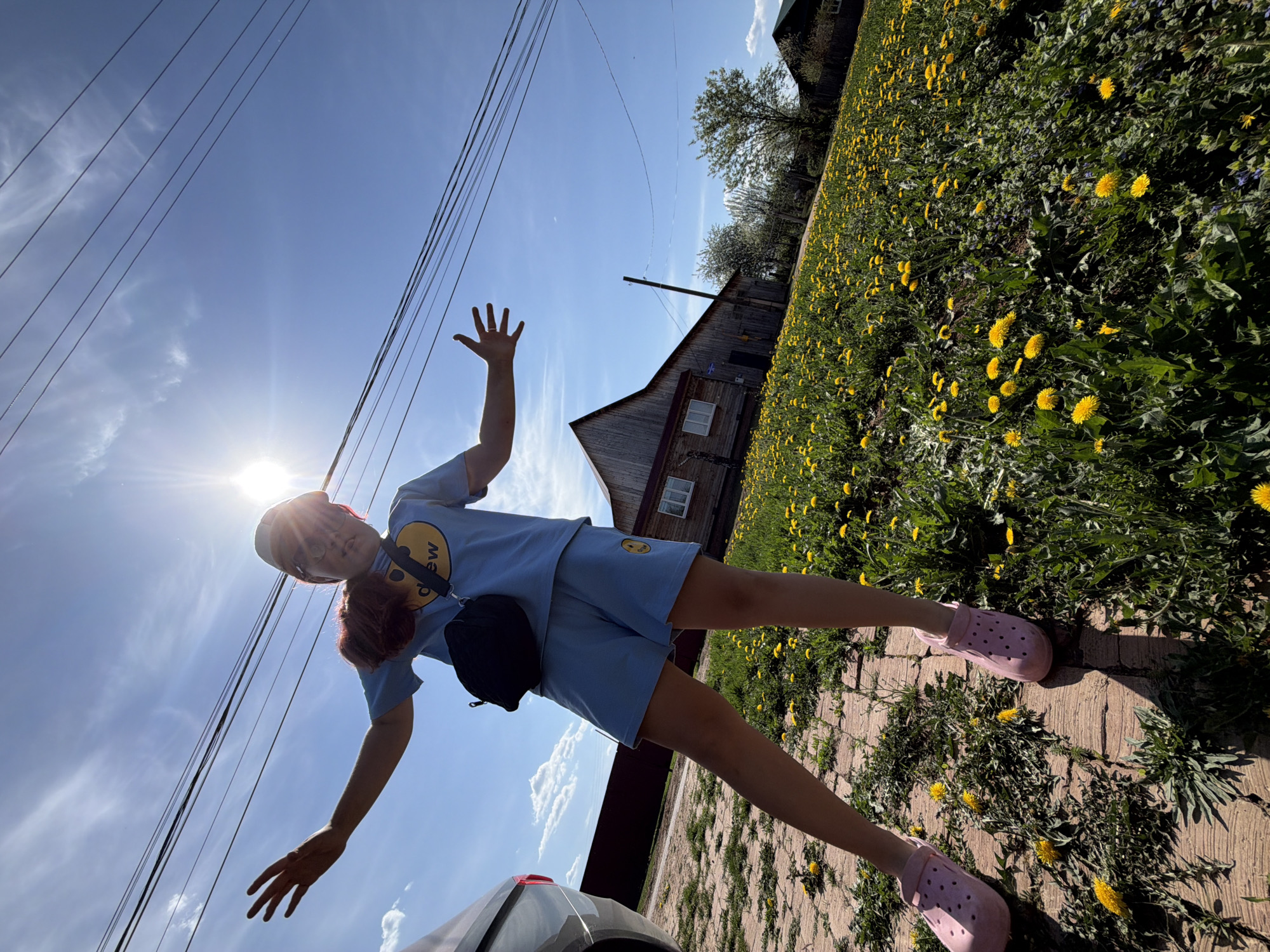A person wearing a light blue t-shirt and shorts with a small black shoulder bag and pink shoes is standing on a brick sidewalk in Чумна, Russia. They have their arms outstretched, with their palms facing forward. To the right, a field of yellow dandelions stretches towards a wooden building with two visible windows and an angled roof. To the left, power lines run across the sky, which is blue with scattered clouds and the sun shining brightly. The front corner of a car is visible in the lower left corner of the image.