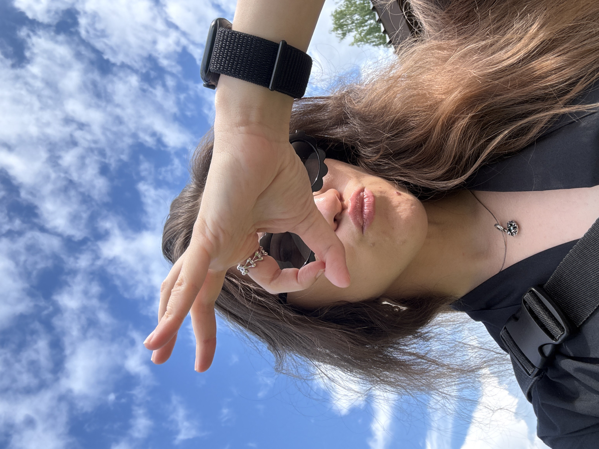 A young woman is captured in a close-up selfie, her face partially obscured by her hand and sunglasses. She wears a black top and a black watch on her left wrist. A silver ring adorns her index finger, and a pendant necklace hangs around her neck. Her long, brown hair is visible, framing her face. The background is a bright blue sky with scattered white clouds, suggesting a sunny day. The image is oriented sideways, with the sky appearing at the bottom and the woman at the top. The scene appears to be outdoors, possibly in a park or open area, given the presence of trees and what looks like a wooden structure in the upper right corner. The overall impression is casual and relaxed, with the woman seemingly enjoying a pleasant day. There is no visible text in the image.