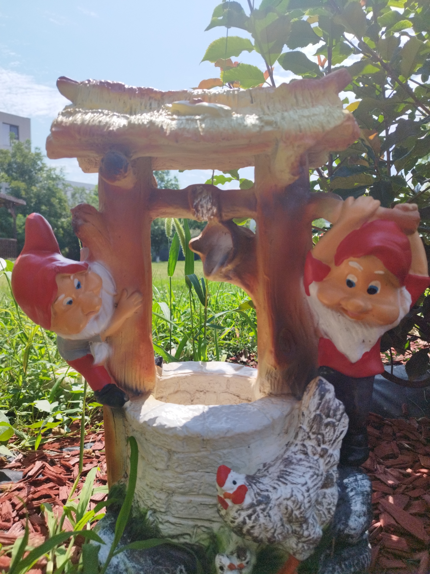 The image depicts a garden ornament featuring two gnomes, a chicken, and a well structure. The gnome on the left is red-hatted and depicted climbing a wooden pillar. The gnome on the right, also red-hatted, is standing with arms raised. A white and grey speckled chicken figurine is positioned in front of the well. The ornament is surrounded by greenery, wood chips, and a grassy area. In the background, a building and trees are visible under a clear blue sky. The image was taken outdoors on a sunny day. There is no information to suggest this scene is located in Bucharest, Romania.