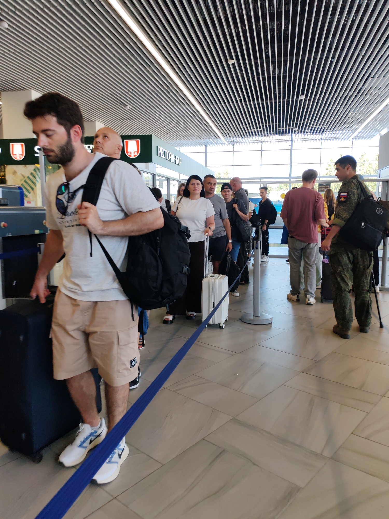 The image depicts a queue of people at what appears to be an airport security checkpoint in Khrabrovo, Russia. A young man with a beard, wearing a light-colored t-shirt, beige cargo shorts, and white sneakers, is in the foreground, holding a dark suitcase and carrying a backpack. He is looking downwards. Behind him, several other individuals are visible, including a man with a bald head, a woman in a white t-shirt, and a person in military camouflage uniform. They are all standing in line, some with luggage. The environment features a modern ceiling with horizontal slats and fluorescent lighting, large windows in the background, and tiled flooring. A blue rope barrier divides the queue. Signage with Cyrillic text, including "РЕСТОРАН-БАР," is present in the background, indicating a service area. The overall scene suggests a typical travel waiting area.
