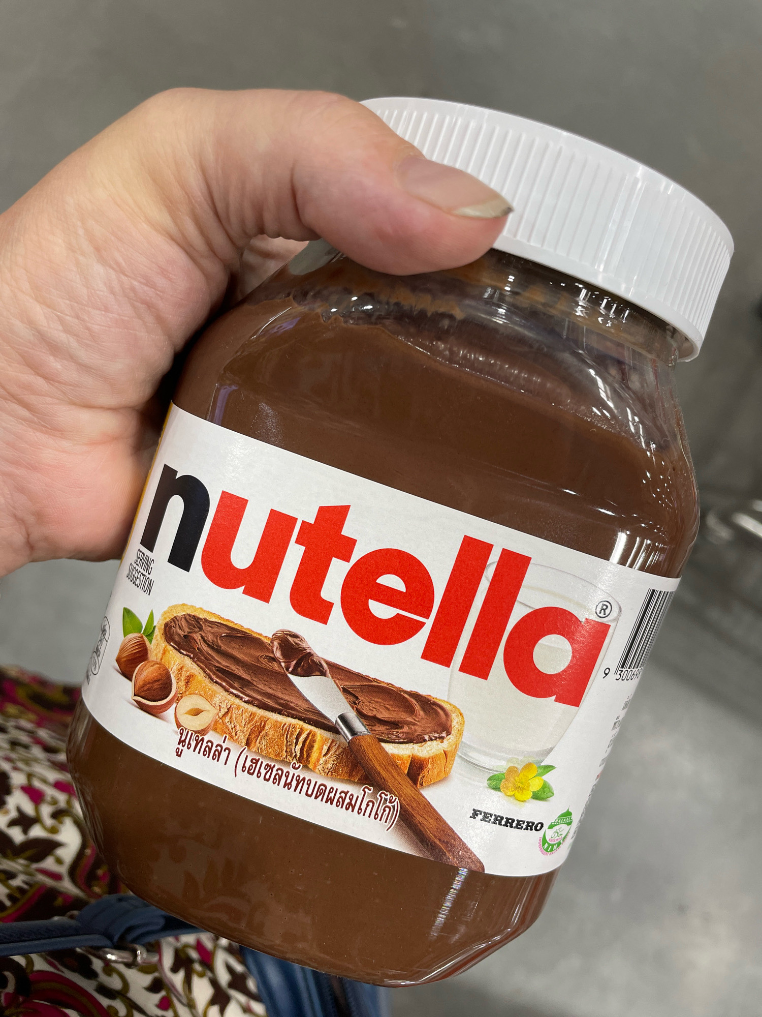A person's hand is holding a large jar of Nutella. The jar has a white lid and a label featuring the Nutella logo, a picture of toast with Nutella and a knife, hazelnuts, a glass of milk, and a yellow flower. The label also contains text in Thai script and the "FERRERO" brand name. The background is blurred and appears to be an indoor setting, possibly a supermarket aisle in Pattaya City, Thailand.