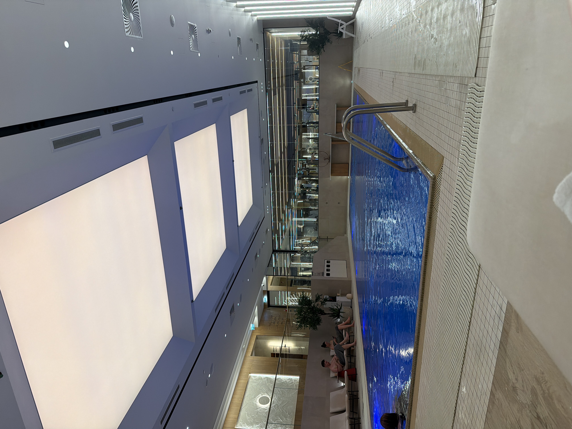A photograph captures a high-angle view of an indoor swimming pool area. The pool, filled with bright blue water, stretches lengthwise along the right side of the frame, complete with a stainless steel ladder entering the water. The pool's edge is lined with tiles, transitioning to a light-colored wooden floor at the very right.
Along the opposite side of the pool are lounge chairs where people are relaxing; one individual is visible in the pool at the bottom. Separating the pool area from an adjacent space, possibly a gym, is a glass wall. Within this space, gym equipment is visible, with some plants placed nearby.
The ceiling, with a linear array of large, rectangular light panels, provides illumination to the area. Vents and other ceiling fixtures are also visible. The image has a modern, clean aesthetic, suggestive of a recreational or wellness facility in a building of Unknown, Unknown.