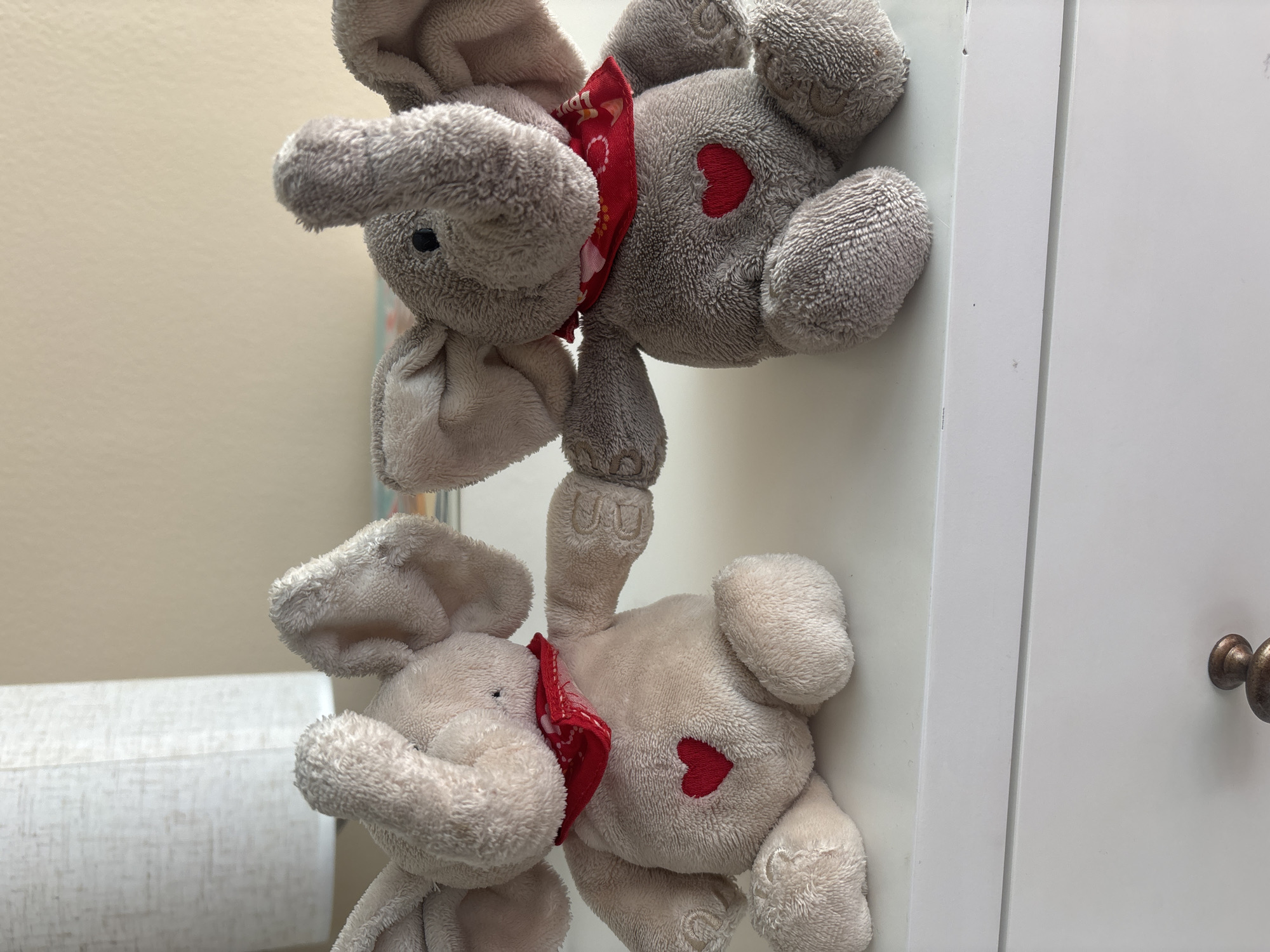 The image shows two stuffed elephants sitting on a white surface, likely a dresser or shelf. The elephant on the left is a light tan color, and the one on the right is gray. Both elephants are wearing red bandanas around their necks with a heart embroidered on their chests. Their front arms are linked, suggesting they are holding hands. Behind them, a portion of a lamp with a textured white shade is visible, and the wall behind is a neutral, light color. The overall scene is domestic and appears to be indoors. There is no indication of the location being Dubai, United Arab Emirates.