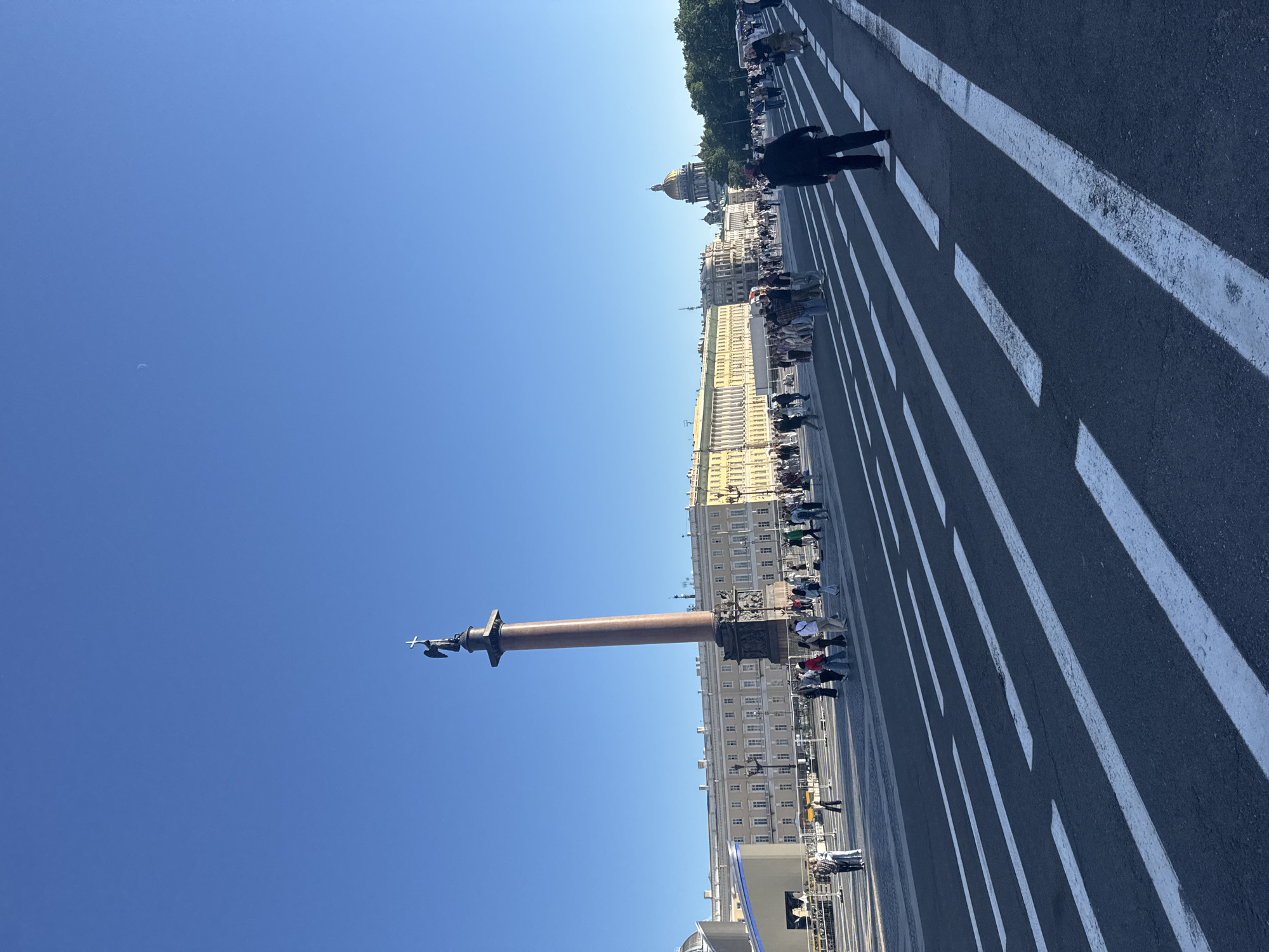 This image captures a sunny day in Saint Petersburg, Russia, with the clear blue sky indicating fair weather. The scene is dominated by a grand architectural ensemble and a prominent monument. A large, tall column, likely a triumphal column, stands in the foreground, topped with an intricate sculpture. In the background, a sprawling building complex with numerous windows stretches along a wide street. Above this, the golden dome of a distant building, possibly a cathedral, is visible against the sky.
The wide street is marked with parallel white lines, suggesting a roadway or a designated pedestrian area. Several people are dispersed along the street, some walking and others gathered, appearing to be tourists or locals enjoying the day. Their presence adds a sense of scale and life to the monumental setting. The perspective of the image is unusual, taken from a low angle and tilted, emphasizing the height of the column and the expanse of the sky. The time of day is clearly daytime, with bright sunlight casting shadows and illuminating the structures. The overall atmosphere seems peaceful and observational.