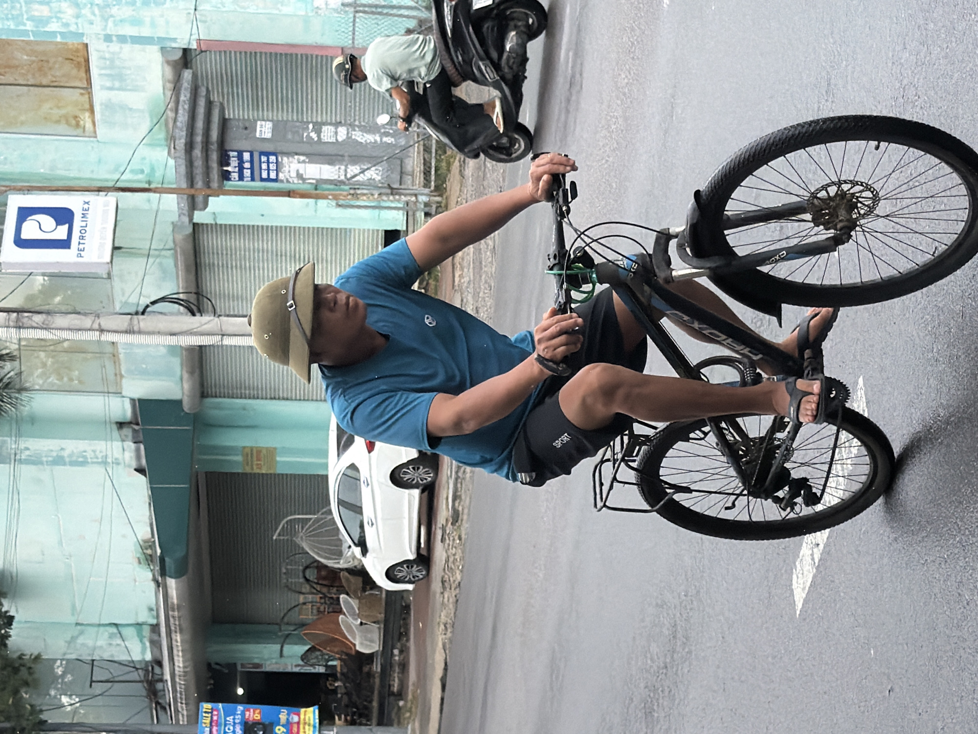 In Nha Trang, Vietnam, on a wet, overcast day, a man wearing a light green helmet and a blue t-shirt rides a black bicycle down a paved street. He is also wearing black shorts with "SPORT" written on the left leg and black flip-flops. The bicycle has a black basket on the rear and a black fender over the front wheel. The man is looking forward as he cycles. Behind him, another person on a scooter is visible. The street appears to be in an urban area with buildings lining the right side of the road. One building has a sign with the logo "P PETROLIMEX" and another sign in Vietnamese. The building on the right is a light blue color with a metallic shuttered entrance. In the background, a white car is parked. The road surface is wet, indicating recent rain.