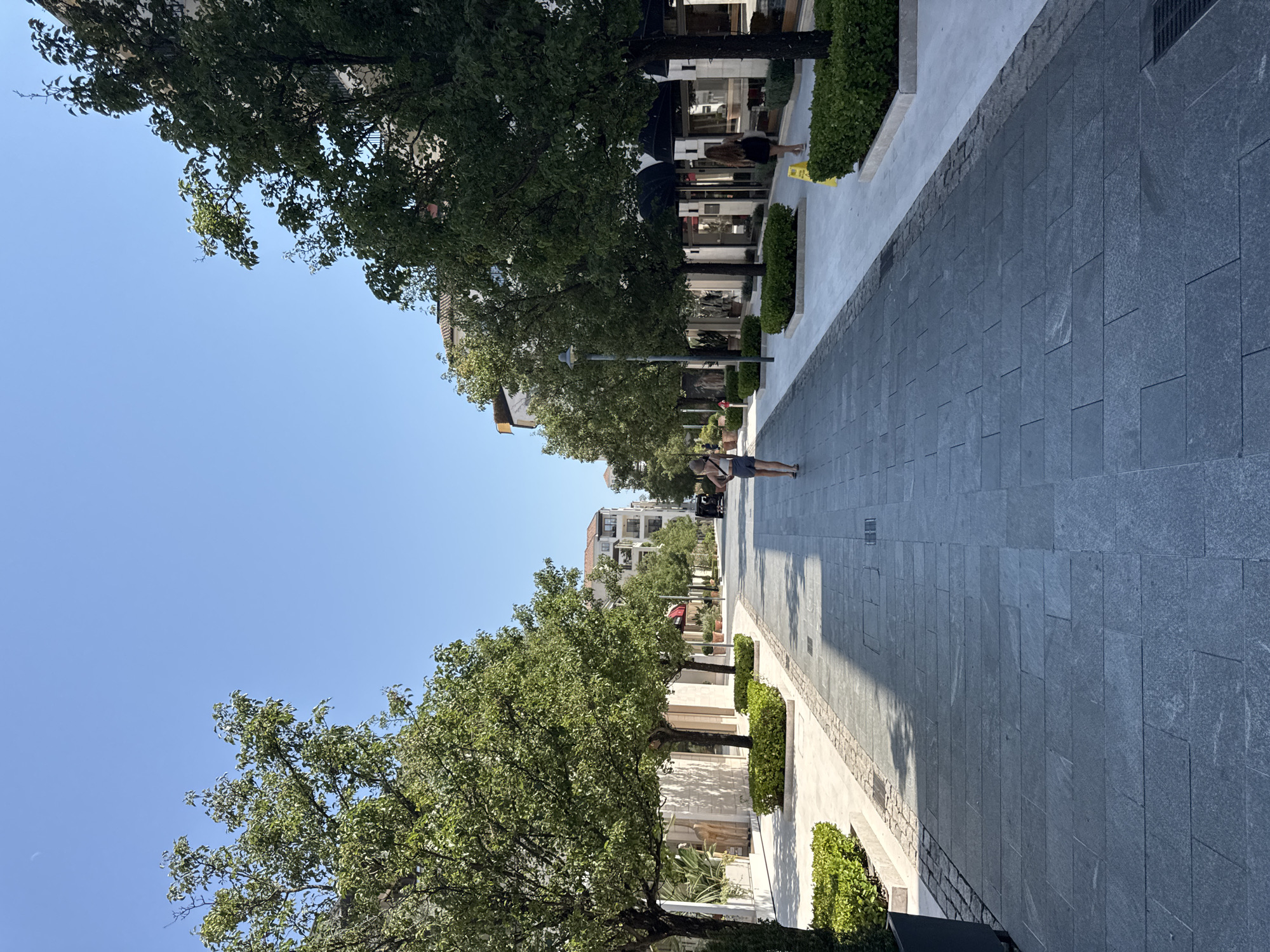 The image depicts a sunny day in Tivat, Montenegro, on a wide pedestrian walkway lined with trees and modern buildings. In the foreground, a woman with blonde hair is walking away from the camera, wearing a light-colored tank top and gray shorts. She is carrying a bag. Further down the walkway, a woman with dark hair is walking away from the camera, wearing a black dress. A black SUV is parked on the right side of the walkway in the distance. The buildings on both sides of the walkway appear to be shops or restaurants, with large glass windows and awnings. The walkway is paved with dark grey stone tiles, and there are neatly trimmed green bushes bordering the path. The sky is clear and blue.