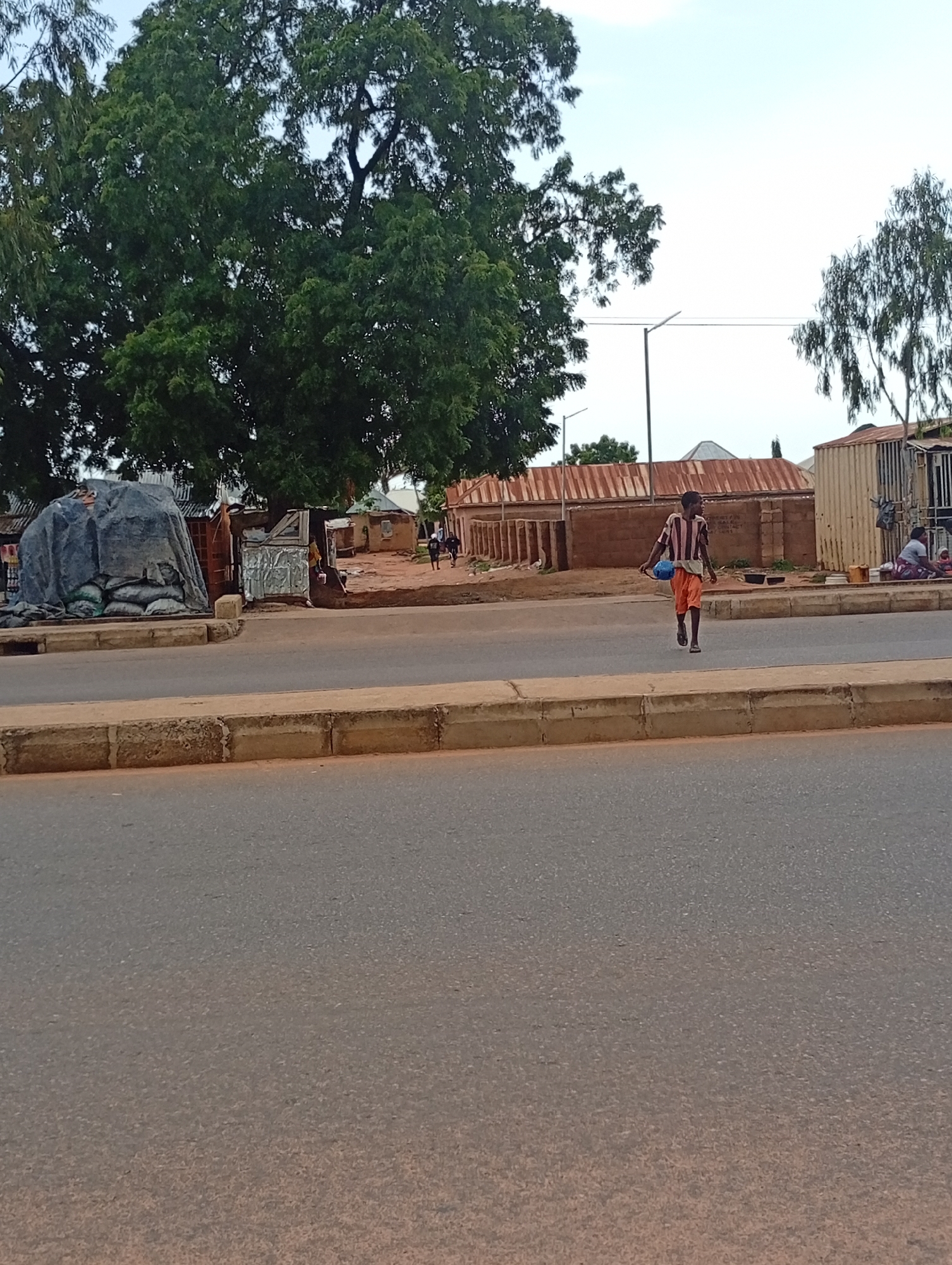 In the city of Wuro Dole, Nigeria, a young boy walks along the side of a dusty road. He is wearing an orange and striped shirt and orange shorts, and carrying a blue object in his right hand. Behind him, several people are visible near a row of small buildings. The environment appears to be semi-arid, with a large tree on the left side of the image and a sparse, distant landscape. A stack of large bags covered in a grey tarp is also present on the left, near the road. The sky is overcast, suggesting it is either early morning or late afternoon.