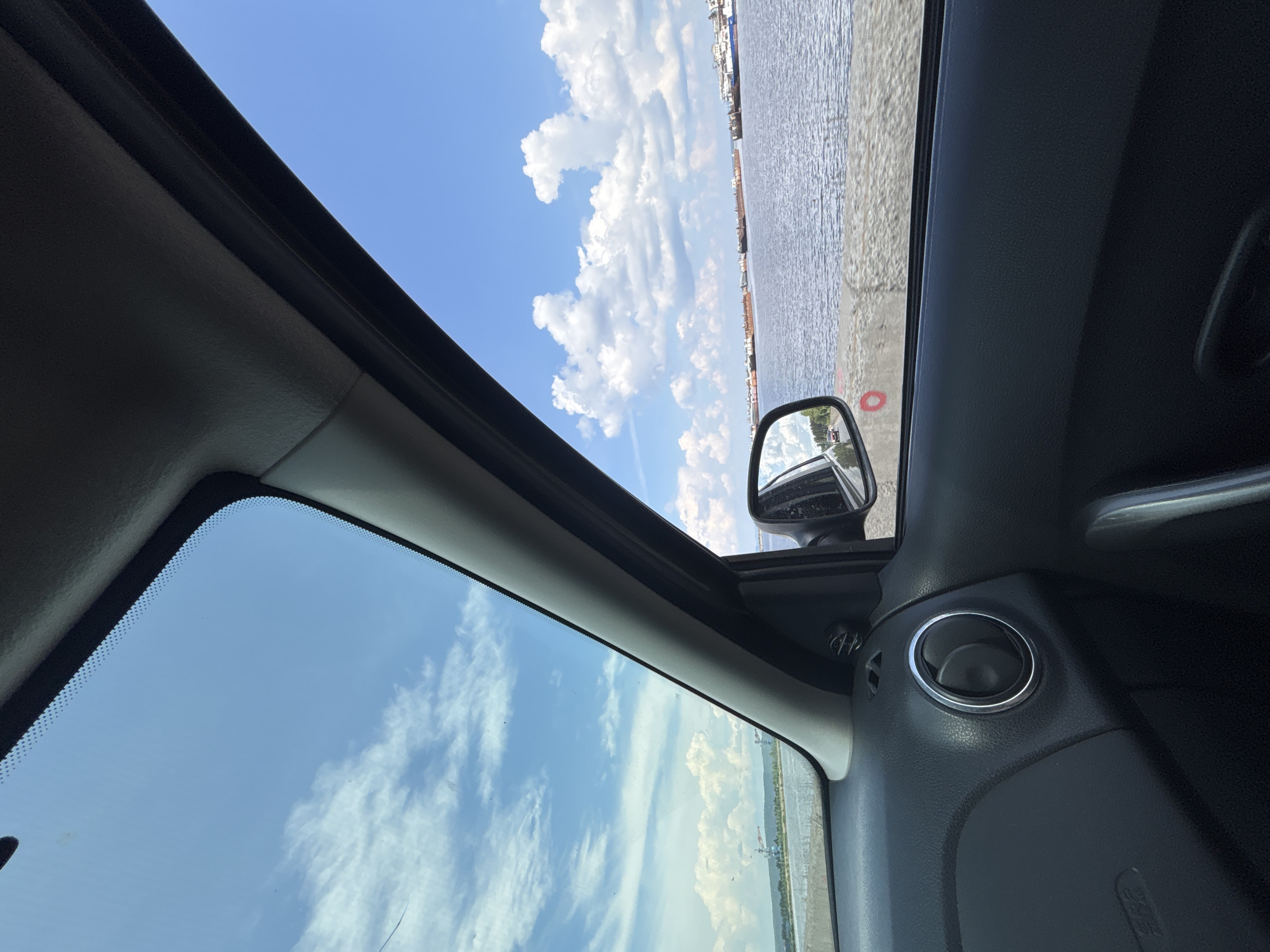 The image is a view from inside a car, looking out the passenger side window and through the windshield. The car appears to be on a road adjacent to a large body of water, likely a river or reservoir. The water is calm and reflects the blue sky and scattered white clouds.
In the distance, across the water, there is a shoreline with some structures that suggest industrial activity or a port. Several ships or barges are visible on the water, some appearing to be docked or anchored. The sky is bright blue with fluffy white clouds, indicating a clear day.
The interior of the car is visible in the foreground, showing the black dashboard, a circular air vent with a chrome ring, and a portion of the door panel with a handle. The side mirror is also visible, reflecting a road with a car driving on it.
The photograph likely depicts a scene in or around Chaikovsky, Russia, a city situated on the Kama River, which is known for its industrial port and water-related activities. The presence of the river, ships, and potential industrial structures aligns with the geographical characteristics of Chaikovsky. No people are directly visible in the image.