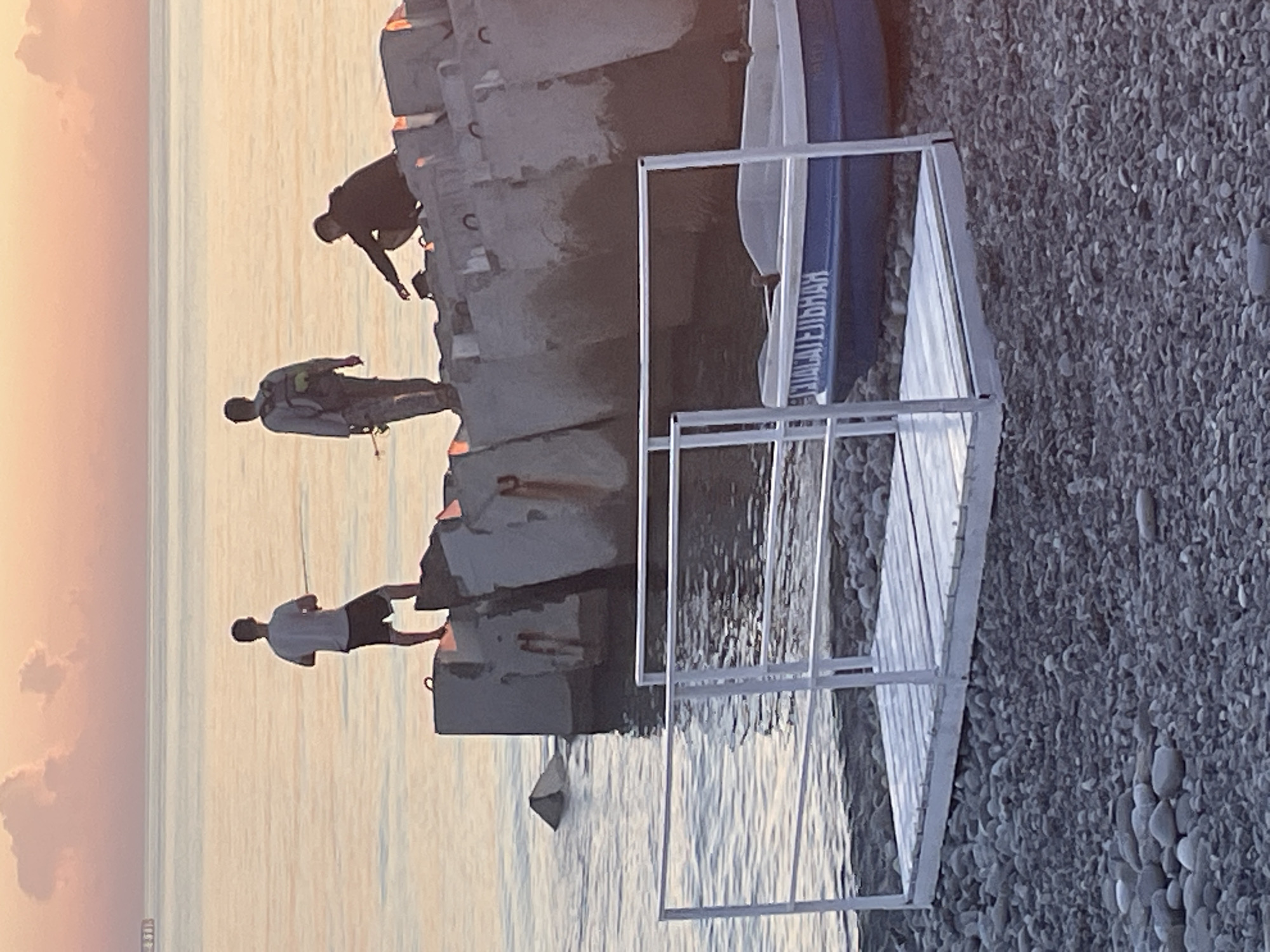 The image shows a beach scene at sunset with three individuals. One person is standing on a concrete structure, fishing. Another person is walking away from the viewer on the same structure. A third person is crouched on the structure, facing the water. A blue boat with white trim is beached on the shore in the foreground, and a small metal ramp with white railings extends from the beach into the water. The sky is painted with hues of pink and orange, and the water is calm. The background consists of the sea and a hazy horizon. The ground in the foreground is covered in pebbles.