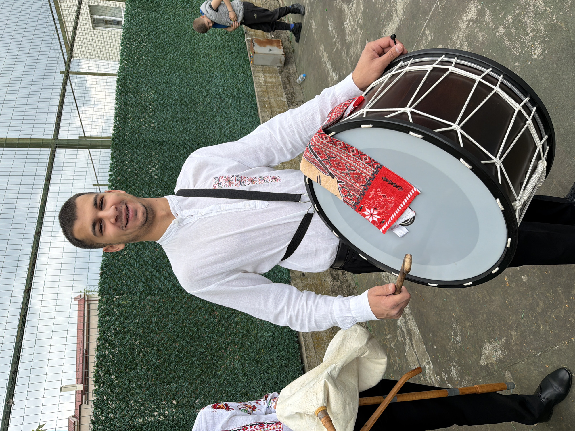 A man wearing a white long-sleeved shirt with traditional red and black embroidery on the left chest stands outside holding a large drum. The drum is black with white rope tensioners, and a red and white embroidered cloth rests on its surface. A black strap goes over the man's left shoulder and supports the drum. The man holds a wooden drumstick in his right hand and is smiling towards the camera. Beside him on the ground are bagpipes.
In the background is a green hedge and a wire fence. Beyond that, parts of buildings are visible. There is a person lying on the ground to the right. The ground appears to be concrete or asphalt. The city of Rakovski, Bulgaria is not mentioned in the image.
