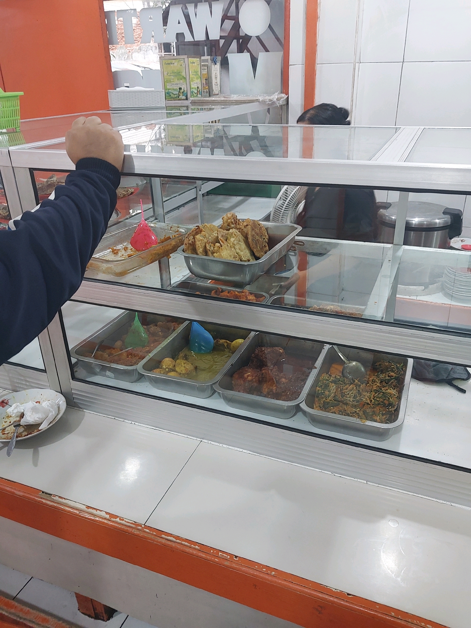 An indoor shot shows a hand reaching into a display case at a food stall in Malang, Indonesia. The display case is made of glass and metal and is filled with various food items in metal trays. Some of the food visible includes fried items, a stew-like dish, and a green vegetable dish. A white plate with leftover food and a napkin is visible in the foreground. In the background, a person's head with dark hair is visible, peeking over the top of the display. The stall appears to be part of a larger establishment with white tiled walls and orange trim. The overall impression is that of a busy food vendor serving customers.