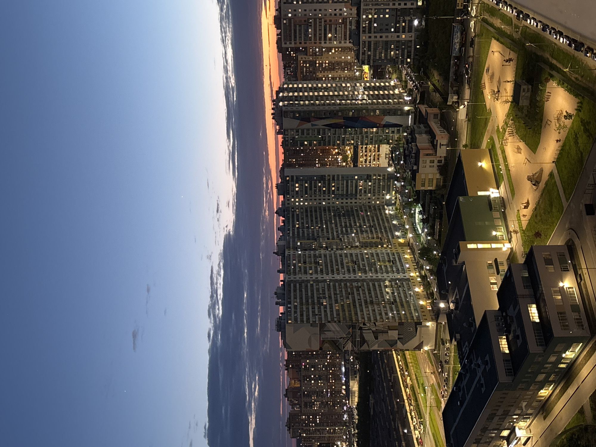 The image depicts an aerial view of a cityscape at dusk. Predominantly, the scene is dominated by tall apartment buildings, many with illuminated windows, suggesting nighttime activity. Between the buildings are illuminated streets with cars and a brightly lit playground. The sky transitions from a deep blue to a strip of orange and pink at the horizon, possibly indicating sunset or sunrise. A dark band of clouds stretches vertically across the left side of the image.
This scene likely captures the urban landscape of a residential area in Saint Petersburg, Russia, characterized by modern high-rise buildings and planned green spaces typical of new developments in the city.