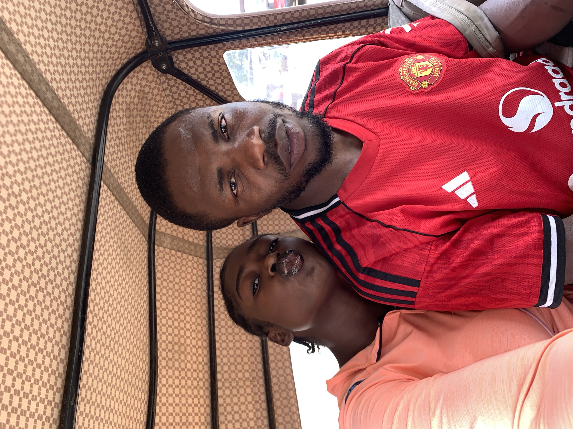 Two individuals are visible within the confines of what appears to be an enclosed vehicle, possibly an auto rickshaw. The background suggests an urban environment.
The first individual, situated at the top of the frame, is a male with short dark hair and a beard. He is wearing a red Manchester United soccer jersey with black stripes on the shoulders and white logos.
The second individual, positioned below the first, is also a male with dark hair styled in braids. He is wearing a peach-colored shirt.
The interior of the vehicle features a patterned beige and brown fabric roof, supported by a black metal frame. The overall image is a close-up shot, capturing the subjects from the chest up. The location is believed to be within the city of Rafin Par, Nigeria.