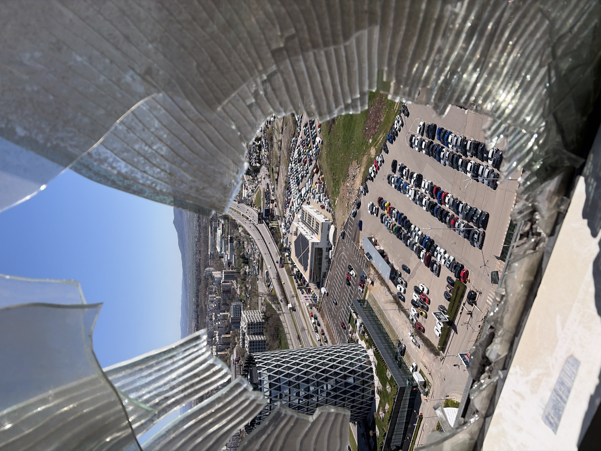 The image shows a view of Sofia, Bulgaria, as seen through broken glass. The cityscape includes buildings, a highway with cars, and a large parking lot filled with vehicles. One prominent building has a distinctive diamond-shaped lattice exterior. The sky is blue. The image is taken from a high vantage point, presumably from a building with damaged or shattered glass. The broken glass fragments are visible in the foreground.