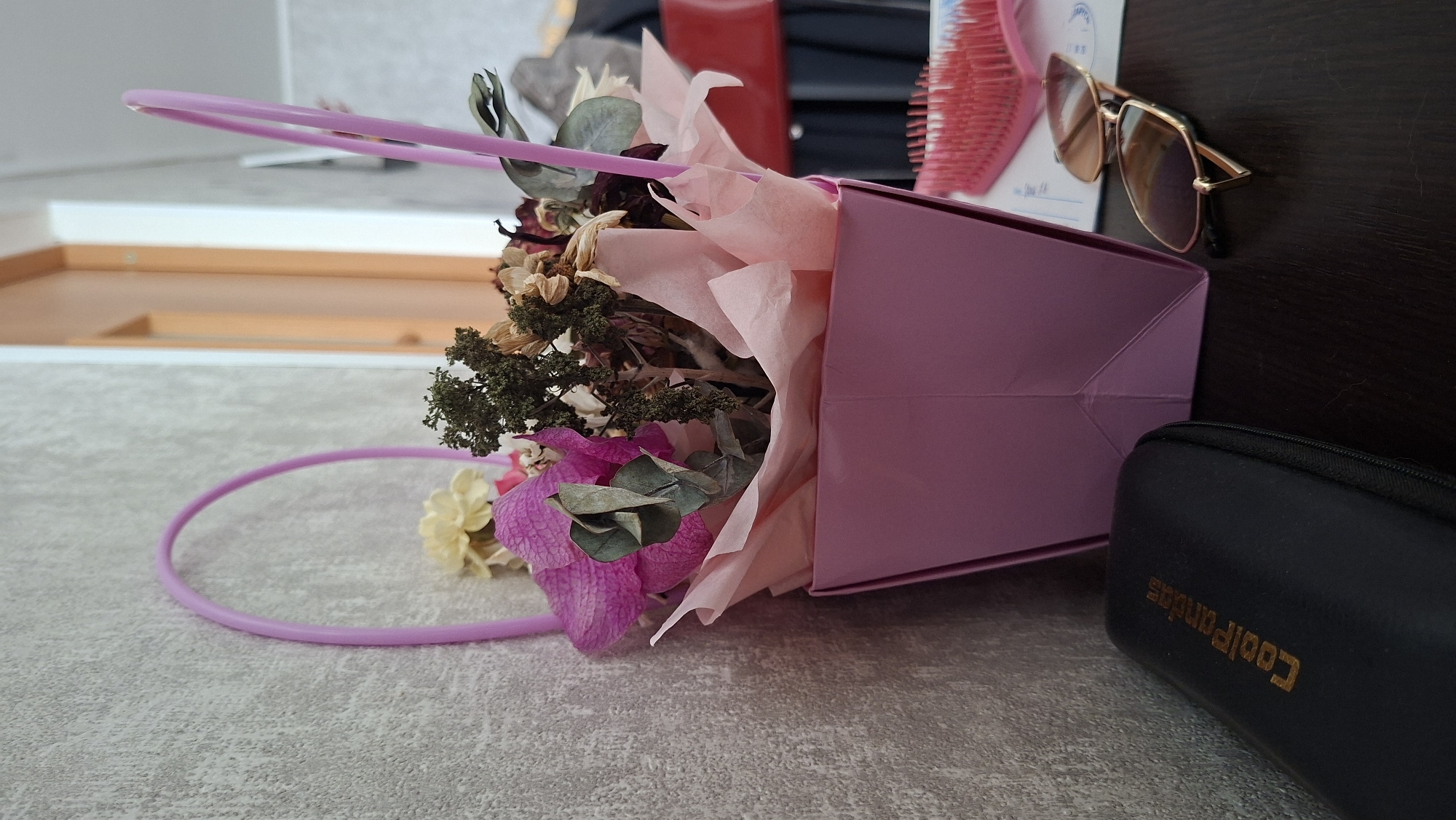 A pink gift basket filled with dried flowers and greenery sits on a dark brown table in front of a textured grey wall. The basket has two pink wire handles. To the left of the basket is a black eyeglasses case with the gold text "CoolPandas" printed on it. To the right of the basket are a pair of gold-rimmed sunglasses with brown lenses, and a pink hairbrush. The scene appears to be indoors. The location is Myadzyel, Belarus.