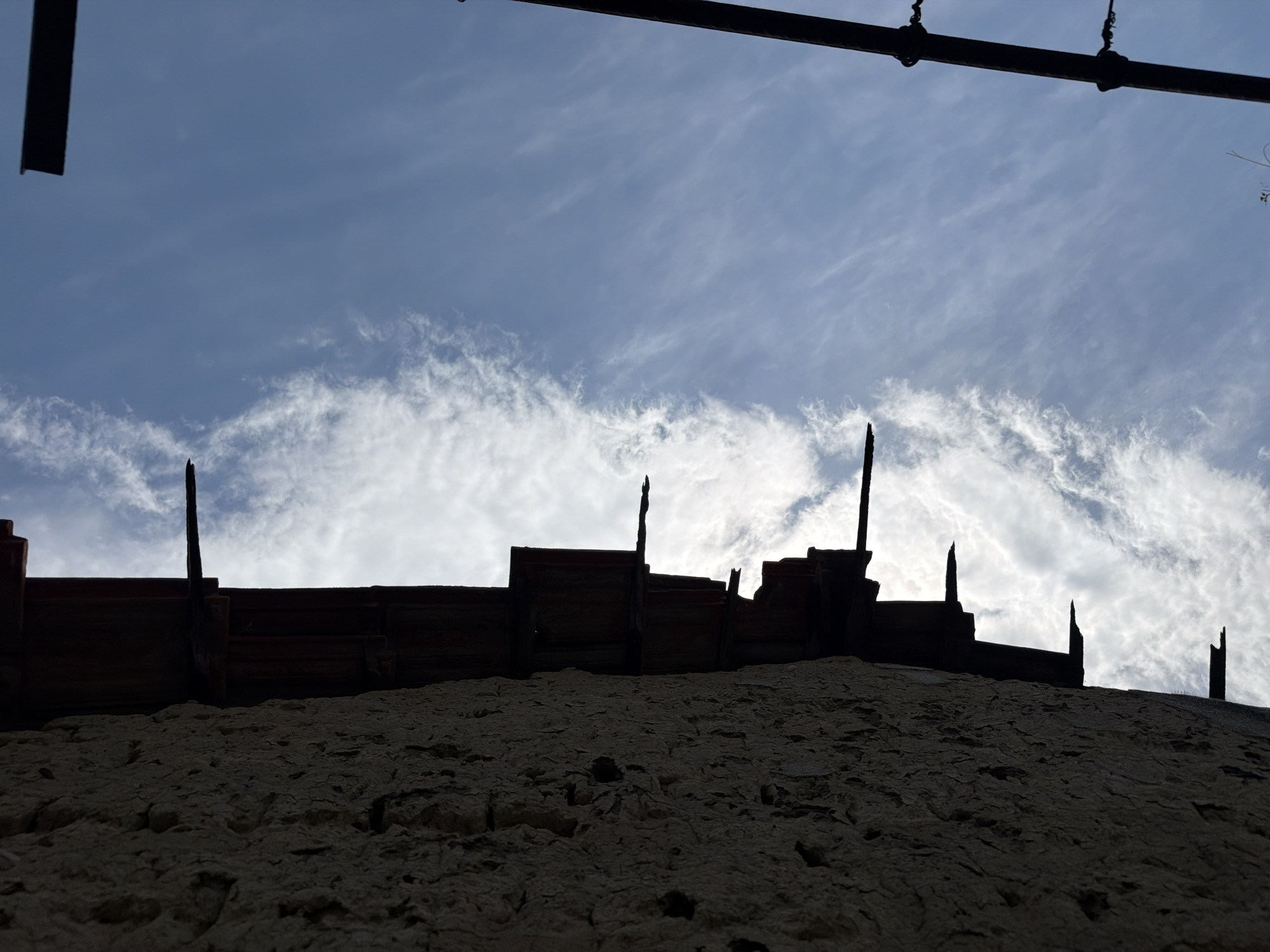 This image, captured in Krumovo, Bulgaria, presents a low-angle, upward-looking view dominated by the silhouette of a building structure against a bright sky. The left approximately two-fifths of the vertical frame is occupied by a rough, textured wall or facade, rendered in deep shadow. This surface exhibits irregular undulations and divots, consistent with aged, possibly concrete or stucco, material.
Extending horizontally from the top edge of this shadowed wall, and partially visible against the sky, are dark, indistinct elements suggesting a roofline or overhang. Several sharp, pointed protrusions, appearing to be broken structural elements like rebar or splintered wood, project from this shadowed area into the illuminated sky. One prominent, slender, dark spike is visible near the upper center of the image, emerging from the dark structure.
The right approximately three-fifths of the frame consists of a clear blue sky, interspersed with scattered, wispy white clouds that exhibit a streaky pattern. In the upper right corner, a dark, rectangular object, potentially a beam or part of a larger structure, is partially visible. Further down and to the right, a vertical dark pole or post is evident, featuring discernible attachments or insulators along its length. The scene is strongly backlit, creating stark silhouettes of the architectural elements against the bright sky. No individuals or active subjects are discernible within the frame.