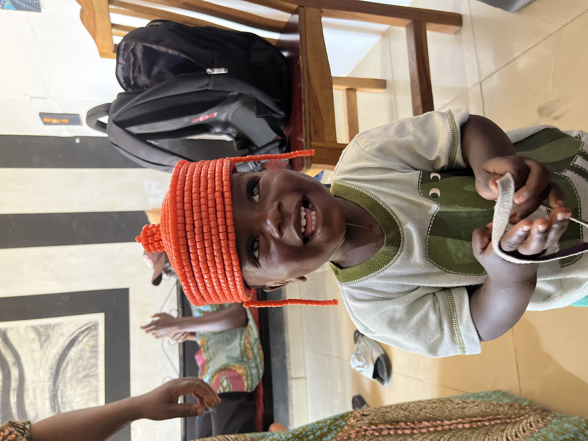 A young Black child wearing a bright orange, beaded hat with hanging strands is smiling at the camera, holding a white object in their hands. The child is wearing a light-colored t-shirt with a green design of eyes. In the background, a black backpack rests on a wooden chair, and a person lies on a patterned blanket on a low surface. To the left, a person's arm and shoulder are visible, wearing a patterned garment. Another person's legs and feet are also visible in the background, wearing a patterned shirt and shorts. The scene appears to be indoors, possibly in a home or community space, with tiled flooring. The general context suggests a gathering or event, potentially in Jos, Nigeria, given the cultural attire.