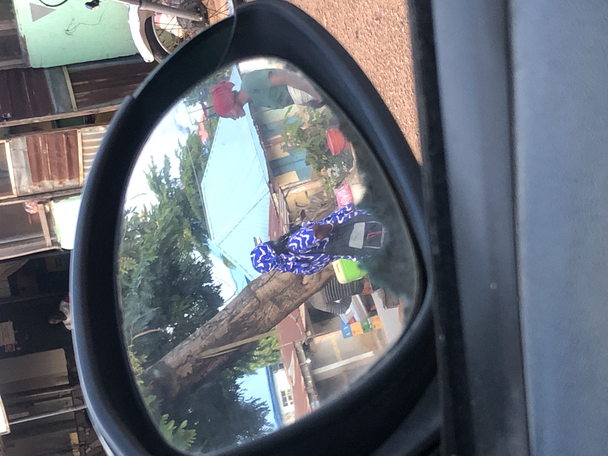 The image is captured from inside a vehicle, looking into its side mirror, which reflects a street scene. The dark frame of the side mirror dominates the central view, with a light grey surface of the vehicle's interior visible below it.
Directly above the mirror, outside the reflection, a dark doorway reveals a person with dark skin wearing a white and red top. To the far right, a portion of a motorcycle, including its front wheel and suspension, is visible against a light green wall.
Within the mirror's reflection:
The scene depicts a sunny, outdoor environment with a reddish-brown dirt or gravel path visible on the right. A prominent large tree trunk stands centrally, with green foliage visible above and around it.
Several rustic buildings are scattered in the background. One structure features a distinct bright light-blue corrugated roof. Other buildings show brown corrugated metal elements and lighter walls. A white building with a darker roof is visible in the distance to the left.
Three individuals are visible within the reflection:
1. **Left-center:** A person with dark skin, facing right, dressed in a blue and white patterned long-sleeved garment and a matching headwrap. They are carrying a dark bag over their left shoulder. A light green bucket or container is positioned near their feet.
2. **Right-center:** A person with dark skin, facing left, wearing a dark green short-sleeved top and a light-colored wrapper around their waist. They have a reddish-orange headwrap. A red bucket is on the ground near their feet. These two individuals appear to be facing each other on the path.
3. **Left-midground:** A partially visible person wearing a grey striped top, standing behind a display of stacked crates in blue, yellow, and green colors.
The overall scene suggests a roadside or market area in a city of Unknown, Unknown, characterized by simple structures and the presence of individuals who might be vendors or passersby.