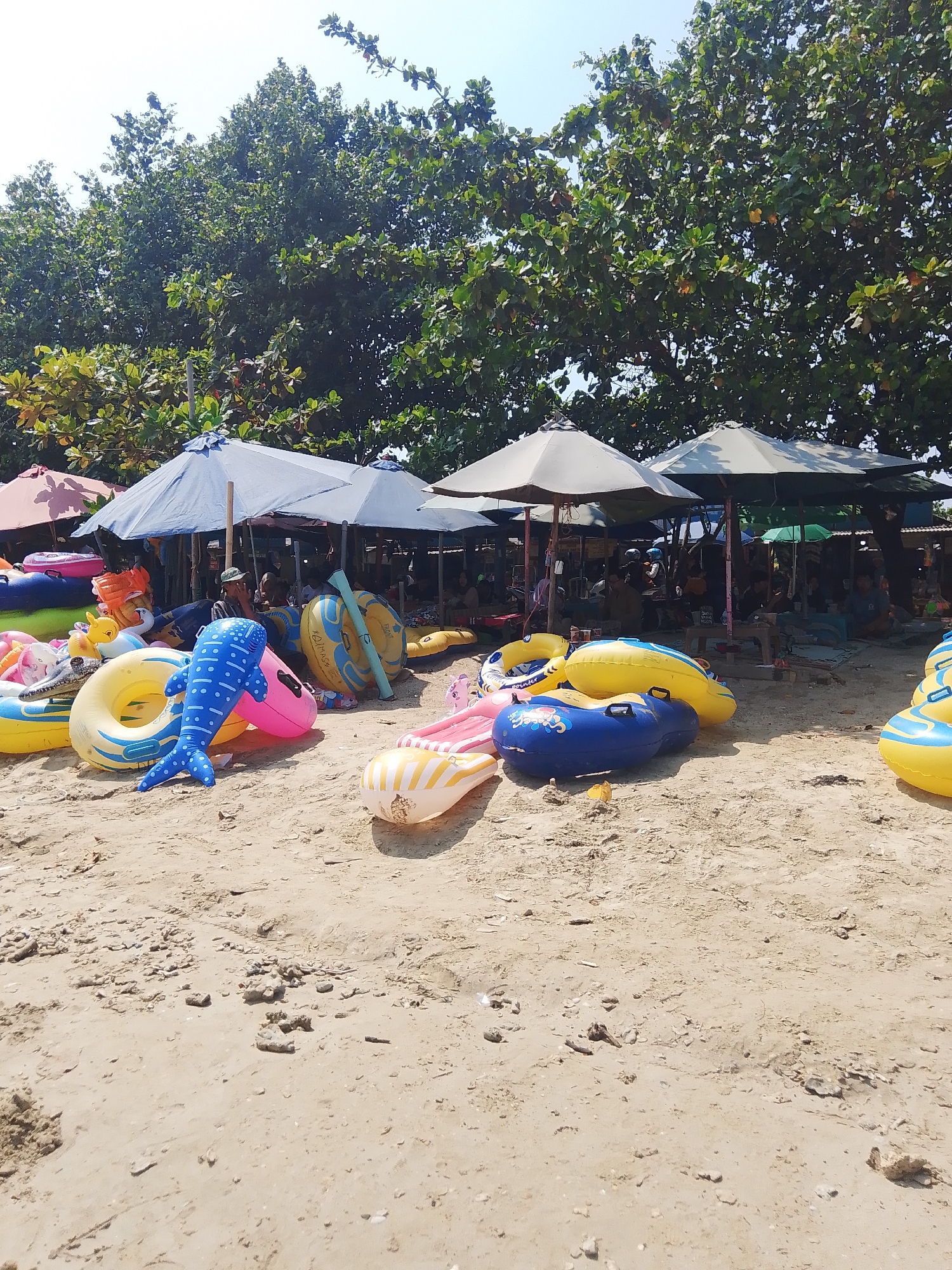 This is a photograph of a beach scene in Pejagan, Indonesia, likely a popular tourist spot, with a focus on inflatable beach toys and shaded areas. In the foreground, the sandy beach is strewn with numerous colorful inflatable rings and pool toys. These include a blue whale-shaped float with white polka dots, pink and yellow inner tubes, and several yellow and blue inflatable rings, some of which are large enough for adults. Some of the inflatables are partially deflated, suggesting they might be available for rent or have been used.
In the mid-ground, several large beach umbrellas provide shade. Underneath these umbrellas, there are glimpses of people sitting or reclining on the sand, indicating a recreational area. Some of these umbrellas appear to be part of a stall or vendor setup, with tables and possibly items for sale visible. The background features lush green trees that create a dense canopy, suggesting a tropical environment. The sky is clear and blue, indicating fair weather. The overall impression is a lively and relaxing beach atmosphere, common in tourist destinations like Pejagan.