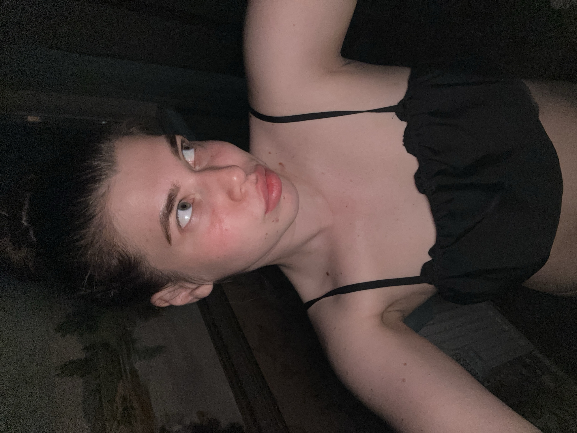 A woman with fair skin and dark hair pulled back is wearing a black top with thin straps. She is lying down, angled to the left, looking directly at the camera. The lighting is dim, casting shadows across her face and body. The background is dark and indistinct, but a patterned, possibly wallpapered, surface is visible in the lower left corner. The scene does not depict any identifiable locations or activities related to the city of Мещёры, Russia.