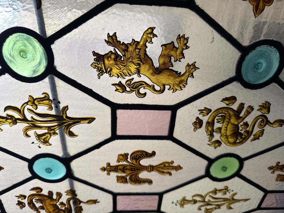 This image presents a detailed close-up of an antique leaded stained-glass window. The window features a geometric pattern of predominantly clear, subtly textured hexagonal glass panes, bordered by thick, dark lead cames. Interspersed within this grid are smaller rectangular panes of light pink glass and circular glass rondels in turquoise-blue and light green, some exhibiting central swirls or bubbles characteristic of hand-blown glass.
Several hexagonal panes are adorned with heraldic and decorative motifs rendered in golden yellow pigment or enamel. Prominent among these are a crowned lion rampant, depicted with forepaws raised and tail curled, facing left; a crowned salamander or dragon, possibly emitting flames, also facing left with a coiled tail; and multiple instances of a stylized fleur-de-lis. Additional motifs include golden floral sprigs with bell-shaped flowers.
The scene focuses exclusively on the architectural and artistic elements of the window, with no individuals, actions, or direct interactions depicted. The light passing through the glass suggests an interior perspective. This specific type of heraldic and decorative stained glass is located within the Musée de Cluny (National Museum of the Middle Ages), situated in the Latin Quarter of Paris, France.
