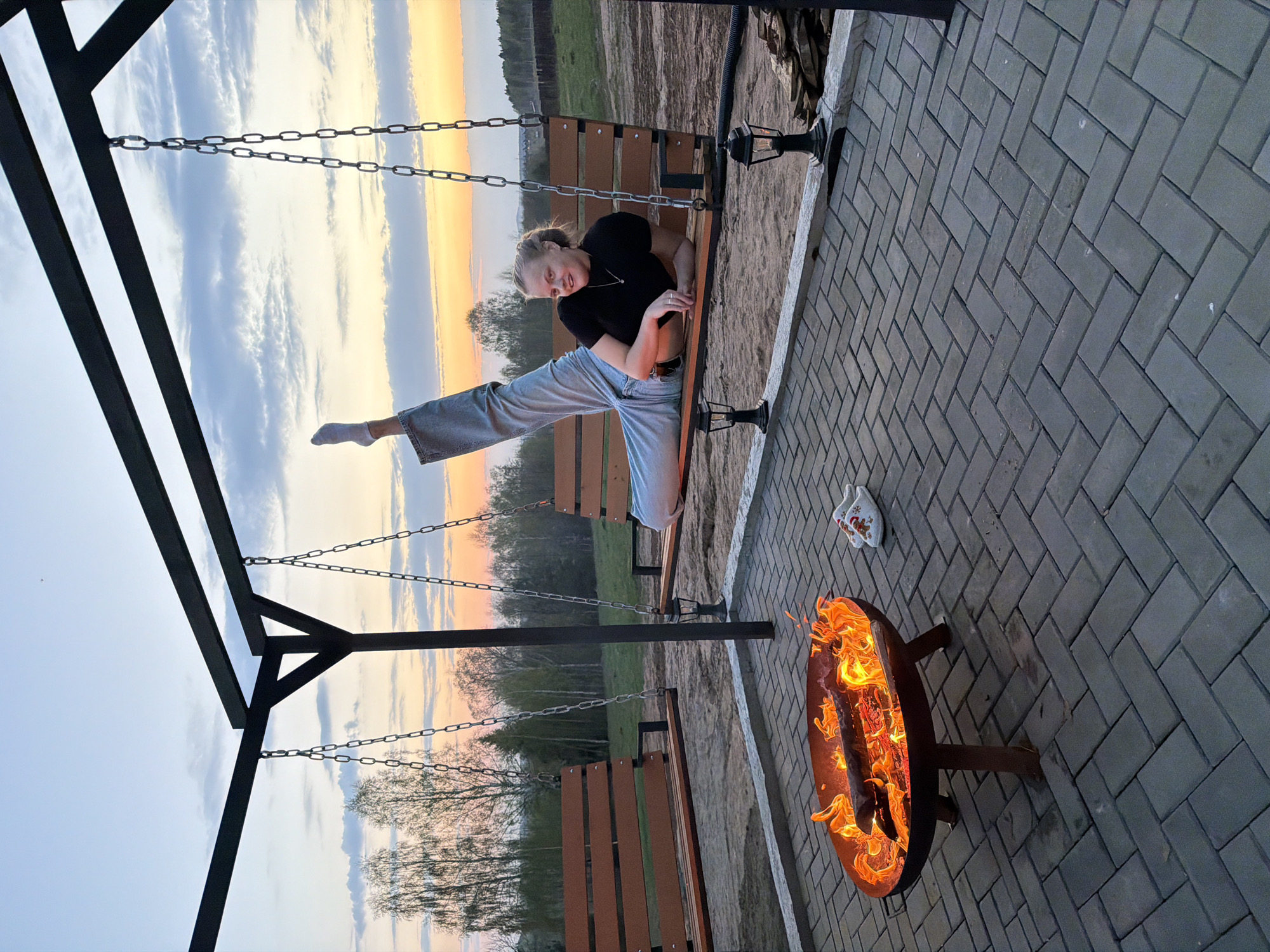A woman is sitting on a swing set with her leg extended straight up, in Kamennoye, Russia. The swing set is made of dark metal and wood. The woman is wearing a black shirt and light blue pants. A fire pit is burning nearby on a brick patio. A pair of white socks and an oven mitt are next to the fire pit. In the background, there is a field and trees, with a colorful sunset sky.