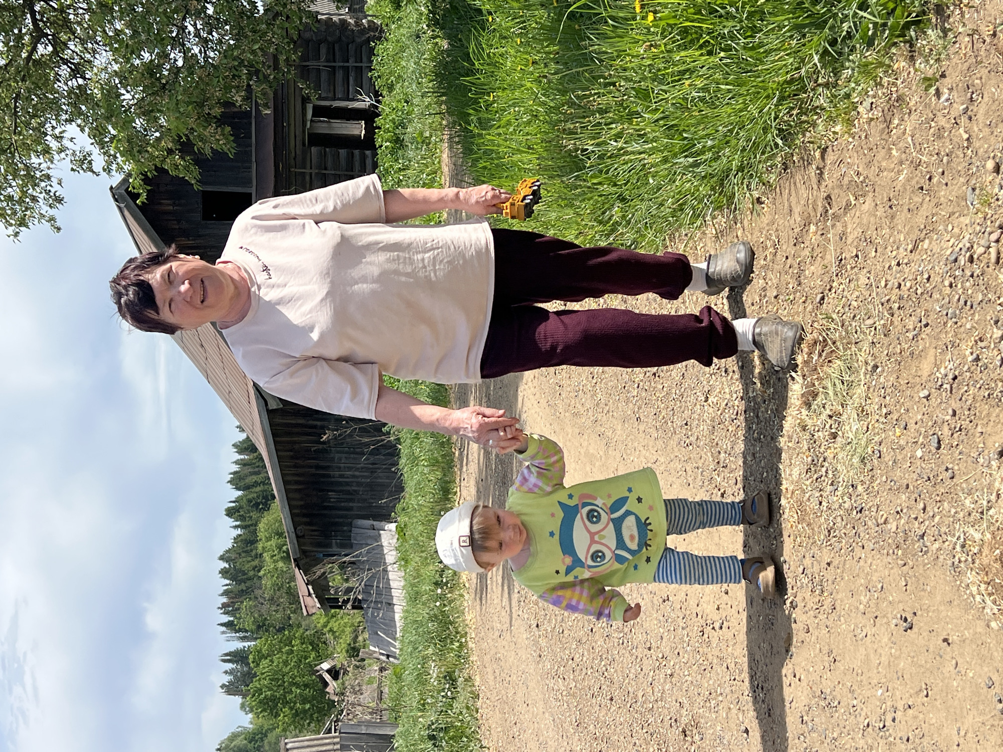 An eye-level, rotated shot shows a woman and a young child walking hand-in-hand on a gravel road near a rustic, dilapidated wooden building. The woman, likely an older adult, wears a light-colored t-shirt, dark pants, and grey shoes. She is smiling, holding a small yellow toy in her free hand. The child wears a white cap, a light green shirt with an owl design, blue and white striped leggings, and dark shoes. The gravel road is lined with grass on one side. The wooden building appears weathered, with some structural elements missing. Behind the building, a forested area with green trees is visible under a partly cloudy sky. The location appears rural, possibly near Panteleevka, Russia, given the style of the building and the surrounding landscape.