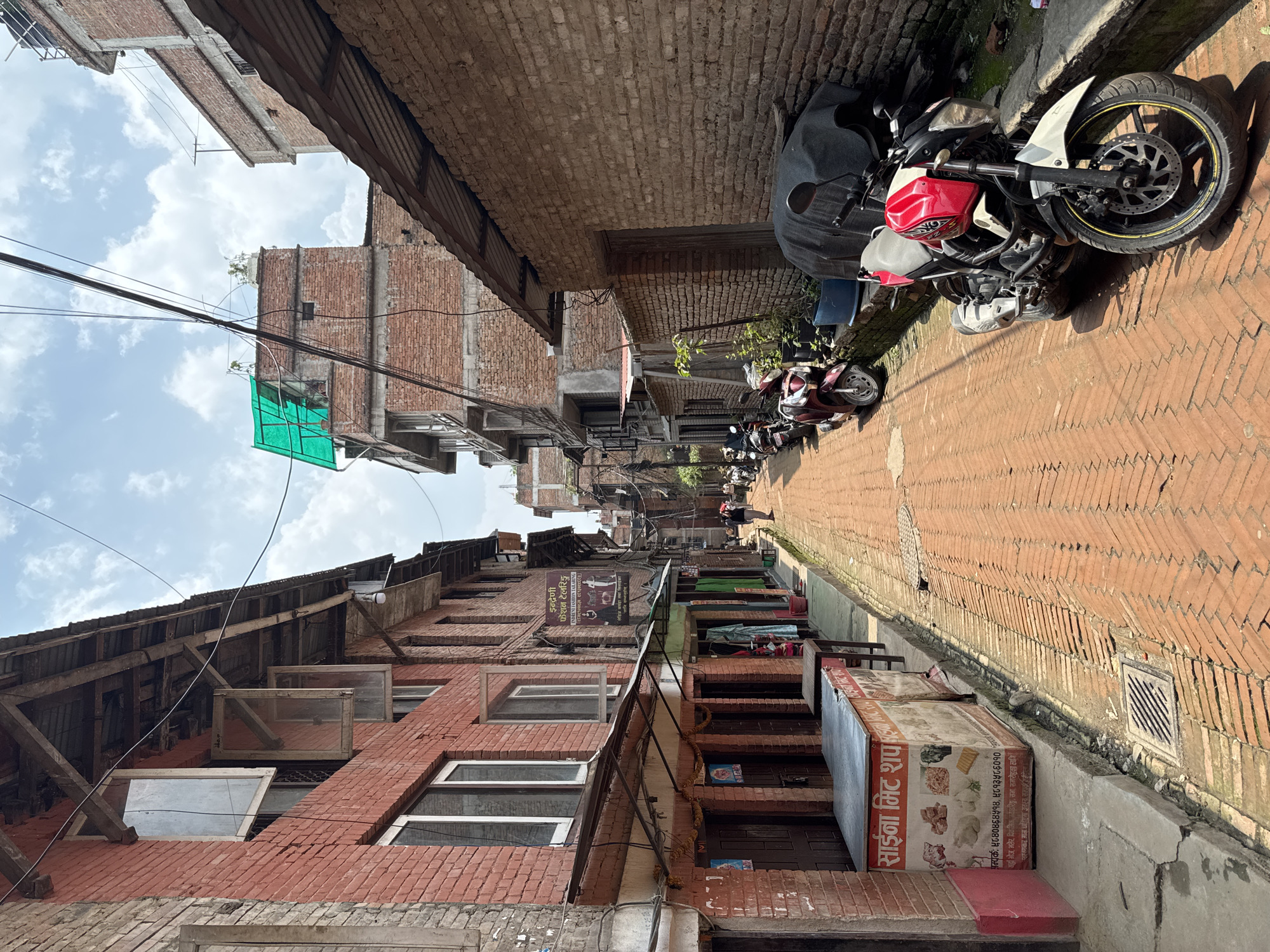 The image shows a narrow, brick-paved street in Bhaktapur, Nepal. Buildings made of red brick line both sides of the street, with traditional wooden windows and overhanging roofs. Power lines are visible overhead. Several motorcycles are parked along the street. A shop sign in Nepali script is visible on the right side. Other commercial signs are also present. In the distance, a person is walking away from the camera.