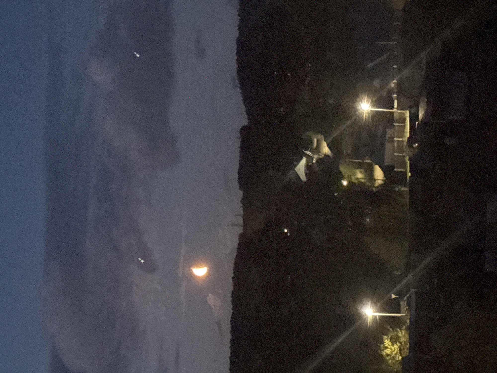 The image presents an outdoor nighttime scene, featuring a partial moon. To the right of the moon, there is a dark, tree-covered area with two streetlights illuminating the surrounding objects. There are a couple buildings visible. Due to the quality of the image, it is not possible to provide any additional information about the location in Chaikovsky, Russia, other than that it appears to be a residential area with streetlights.