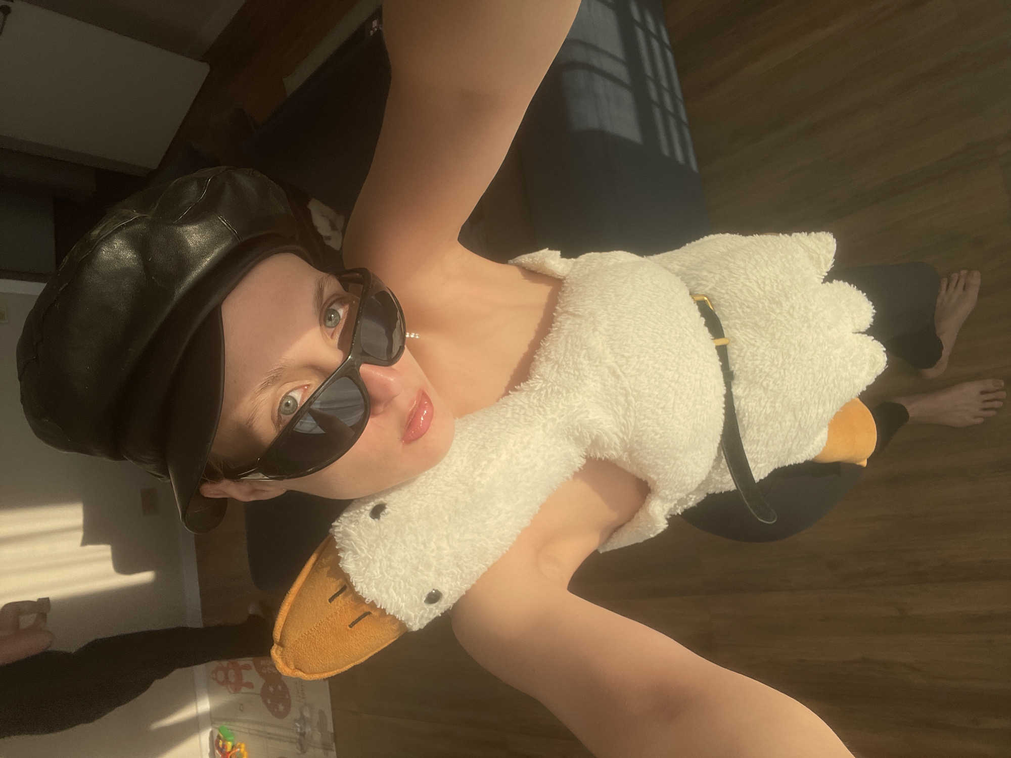 A person wearing a black beret and sunglasses is holding a plush white goose. The person is standing indoors on a wooden floor, with a bed or sofa in the background and sunlight streaming through a window, casting shadows. Part of a baby's play area is visible in the lower left corner of the frame. The location is not discernible from the image.