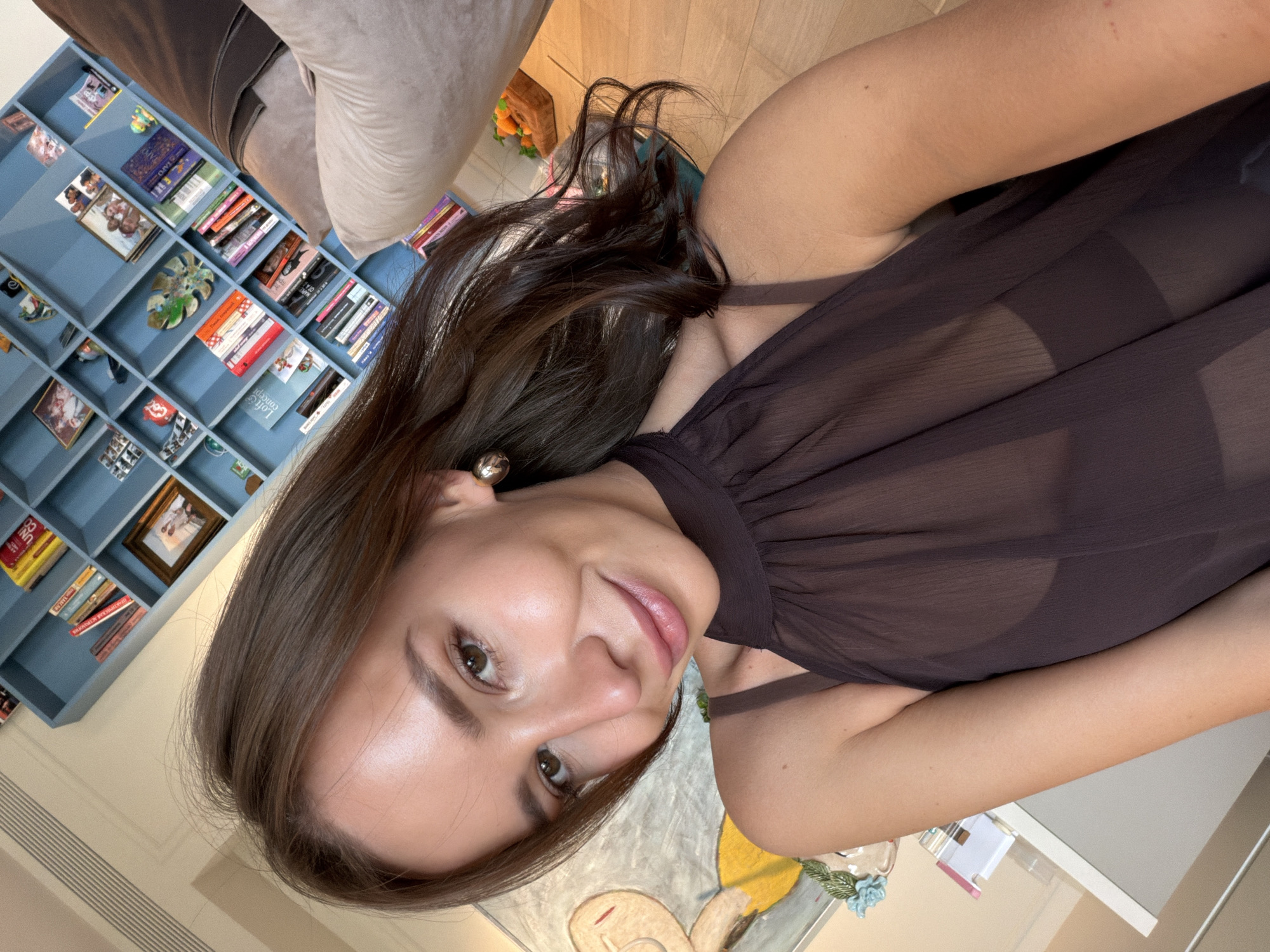 A woman with brown hair is lying on a light-colored couch, wearing a brown sheer top. She is smiling and looking at the camera. Behind her is a blue bookshelf filled with books and other objects. The woman is positioned in a room that appears to be in a residential setting. The location is unknown, and I cannot identify if the room is in Moscow, Russia.