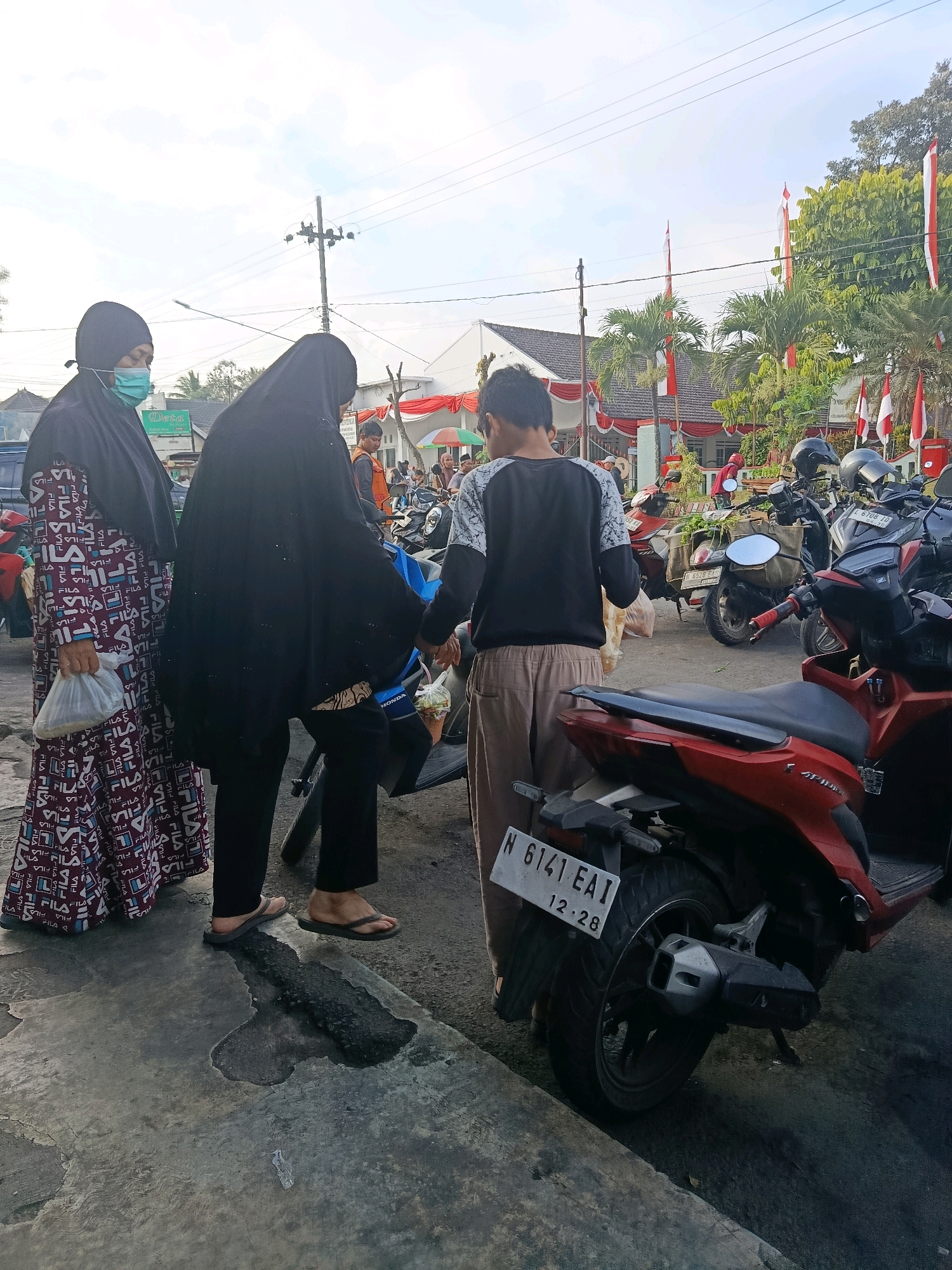 In Bululawang, Indonesia, on a sidewalk next to a street, two women in hijabs and a young man in casual clothing are gathered around a motorcycle. One woman on the left, wearing a mask, holds a white plastic bag. The woman in the middle, dressed in black, is interacting with the young man. The young man, in a black and white long-sleeved shirt and light brown pants, holds a clear plastic bag containing food items. Another motorcycle is parked in the foreground. In the background, a building is decorated with red and white Indonesian flags. Several other people and motorcycles are visible further down the street, suggesting a public gathering or market area. The sky is overcast.