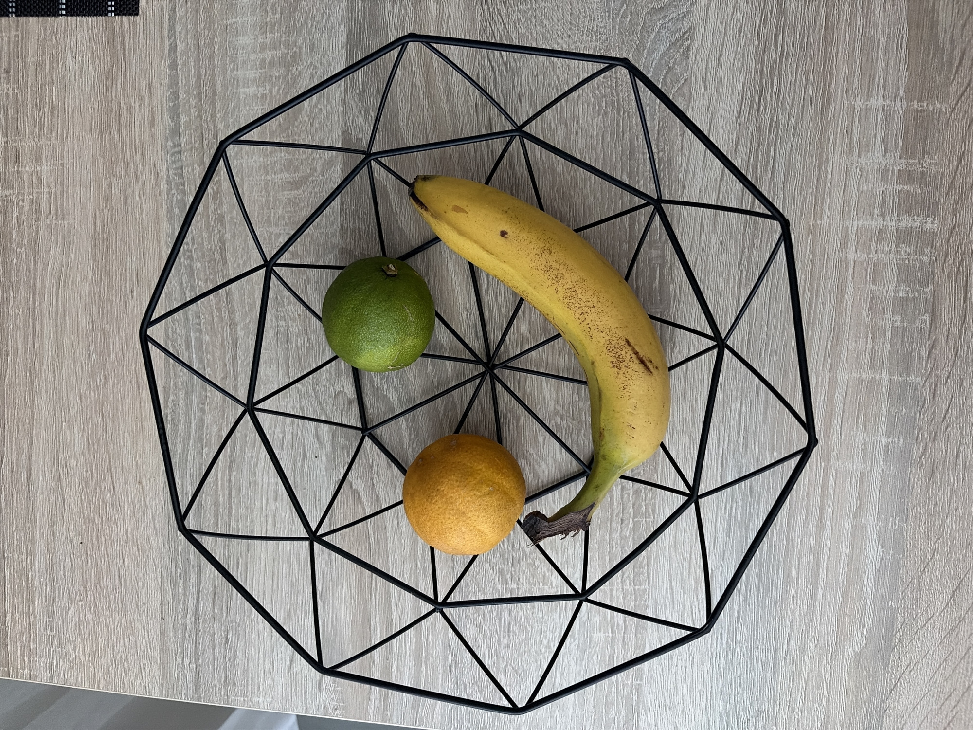 A black geometric wire fruit bowl is placed on a light wood-grained surface. Inside the bowl, arranged to resemble a smiley face, are three pieces of fruit. A yellow banana forms the smile, positioned horizontally across the bottom. To the left of the banana, a small, bumpy, orange-colored citrus fruit, possibly a clementine or small orange, represents one eye. To the right of the banana and slightly above it, a round, green lime serves as the other eye. The surface appears to be a tabletop. The scene is captured from a top-down perspective. There is no indication of the city of Nitra, Slovakia in the image.