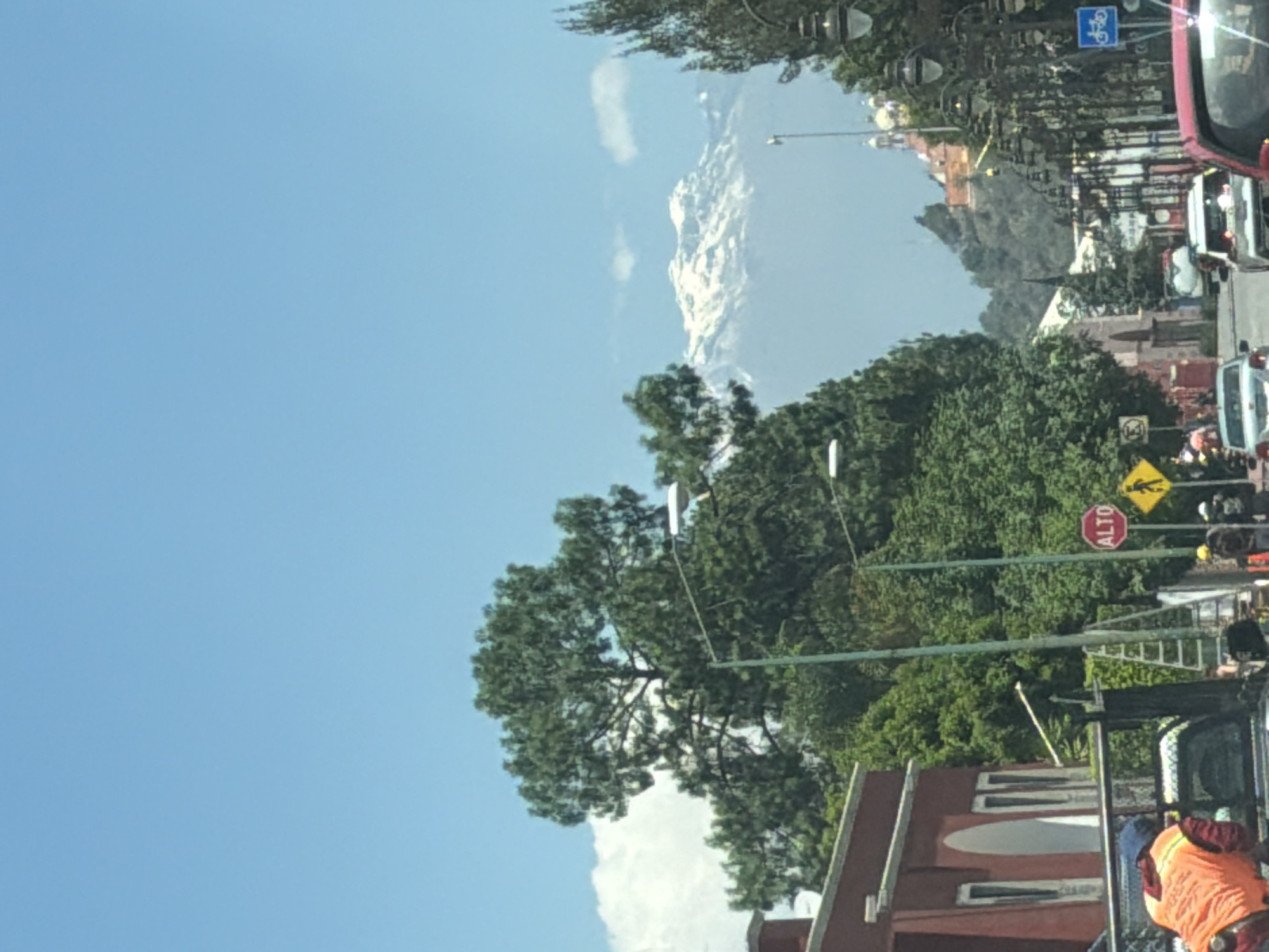 The image shows a street scene in San Andrés Cholula, Mexico, with a view of a snow-capped mountain in the distance under a clear blue sky. The street is lined with trees, lampposts, and buildings. Traffic is visible, including a red vehicle and other cars and trucks. A pedestrian crossing sign and a red "ALTO" (stop) sign are present. A construction worker wearing an orange vest stands near a truck in the foreground.