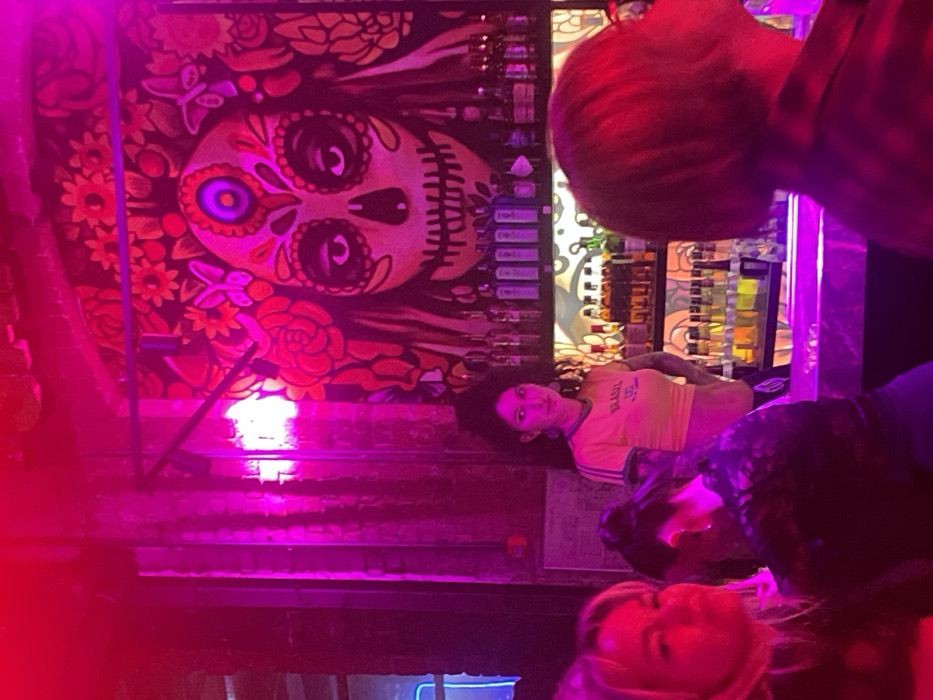 The scene depicts the interior of a bar or club, located in Moscow, Russia, characterized by dominant pink and magenta lighting. A large, ornate mural of a "Sugar Skull" (Calavera) is painted on the back wall, featuring decorative patterns around the eyes, forehead, and mouth, surrounded by flowers and butterflies.
Behind a reflective bar counter, a female bartender with dark, curly hair stands centrally. She is wearing a yellow, short-sleeved top with "BRASIL" visible on the front and displays tattoos on her forearms. She is looking slightly to her left. Behind her, multiple shelves are stocked with various bottles of alcoholic beverages.
In the foreground, several patrons are visible. On the far left, a person with blonde hair is partially seen, facing slightly to the right. In the mid-ground, another person with dark hair tied up is seen from behind, wearing a dark, possibly patterned top. On the right, a third individual with short, reddish-brown hair is viewed from the back, positioned at the bar counter, looking upwards and to the left. Exposed brickwork is visible on the walls, contributing to the establishment's aesthetic.