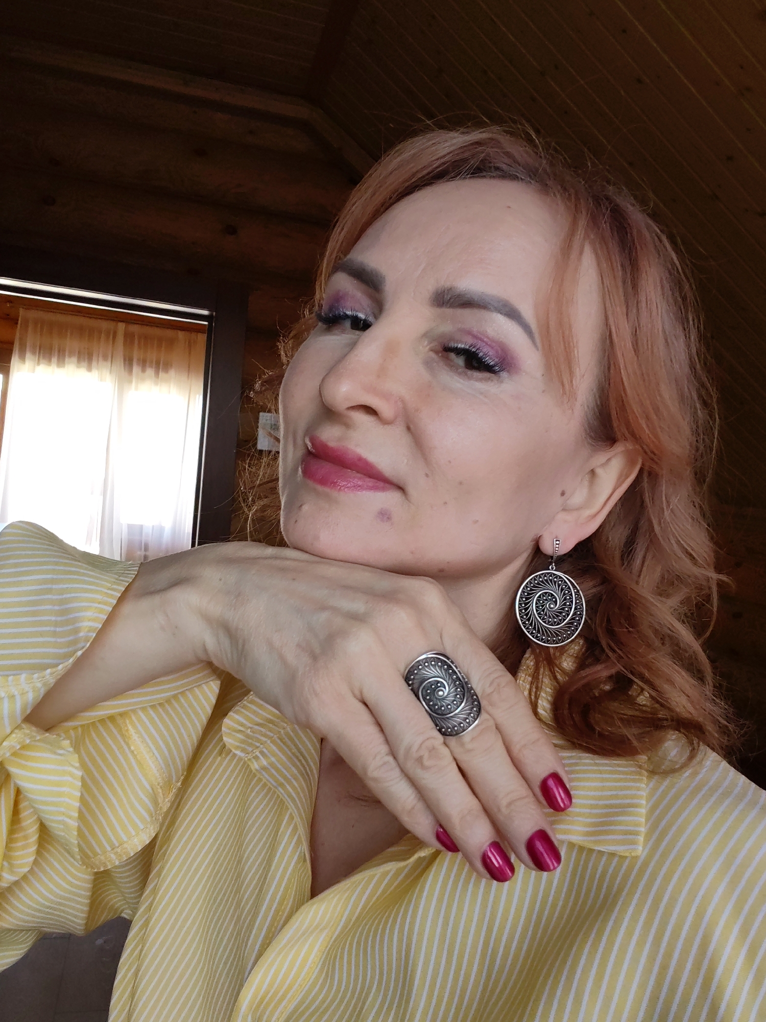 A medium-close shot of a fair-skinned woman with red-blonde hair, wearing a yellow and white striped blouse, silver spiral-patterned earrings, and a matching ring. She is resting her chin on her hand, and she is wearing pink lipstick and purple eyeshadow. The background appears to be a rustic interior with wood paneling and a window with partially drawn curtains. The location is Старое Михайловское, Russia.
