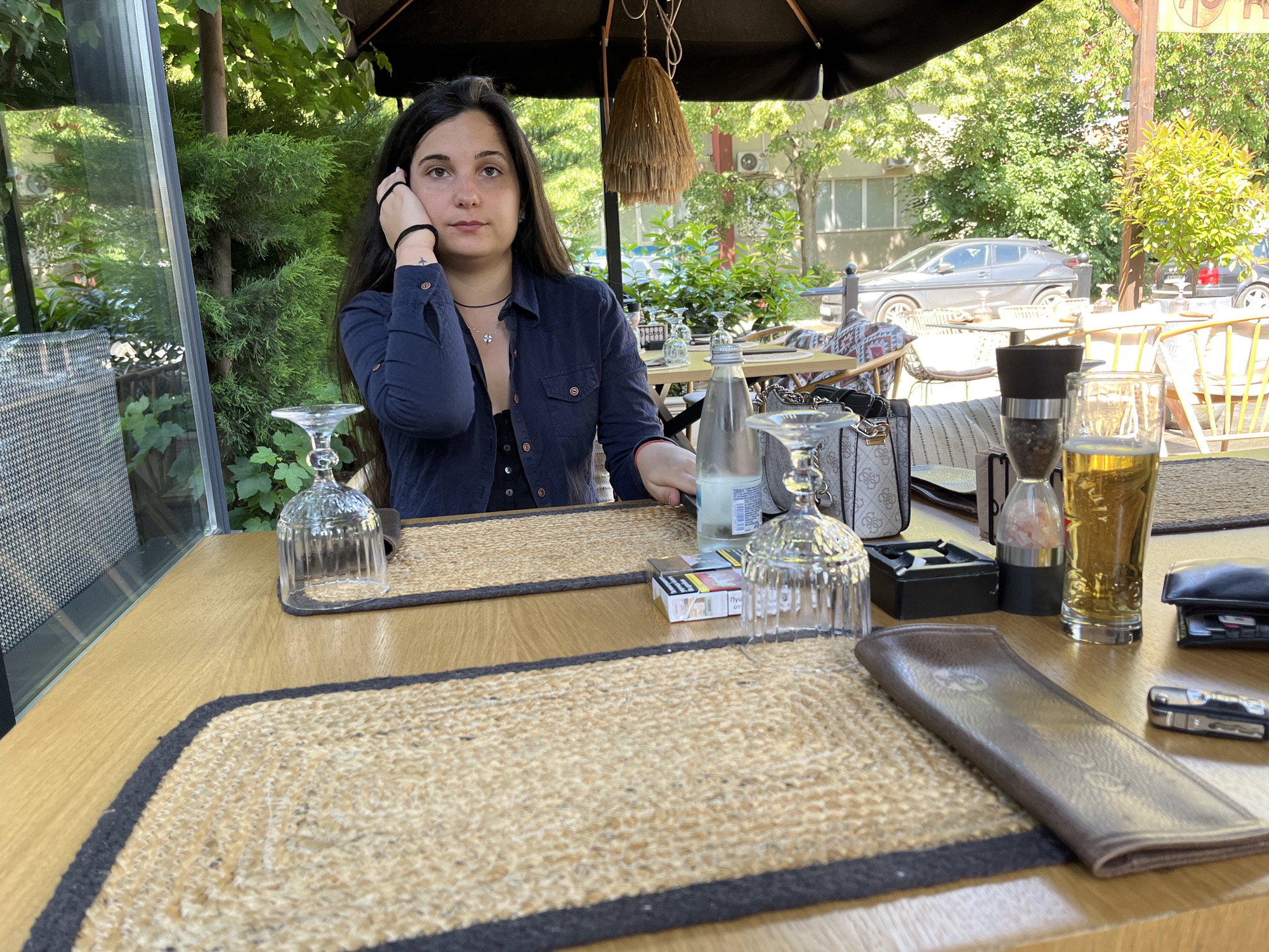 A young woman with dark hair, wearing a dark blue button-down shirt over a black top, is seated at an outdoor table at a cafe. She is touching her ear with her right hand and looking to her left. The table is set with two woven placemats, a glass of beer, a bottle of water, a pack of cigarettes, an ashtray, a salt grinder, and two empty wine glasses. The cafe appears to be located in Sofia, Bulgaria, with lush green trees and buildings visible in the background, suggesting a daytime setting. The table itself is made of light brown wood, and the overall atmosphere is relaxed and informal.