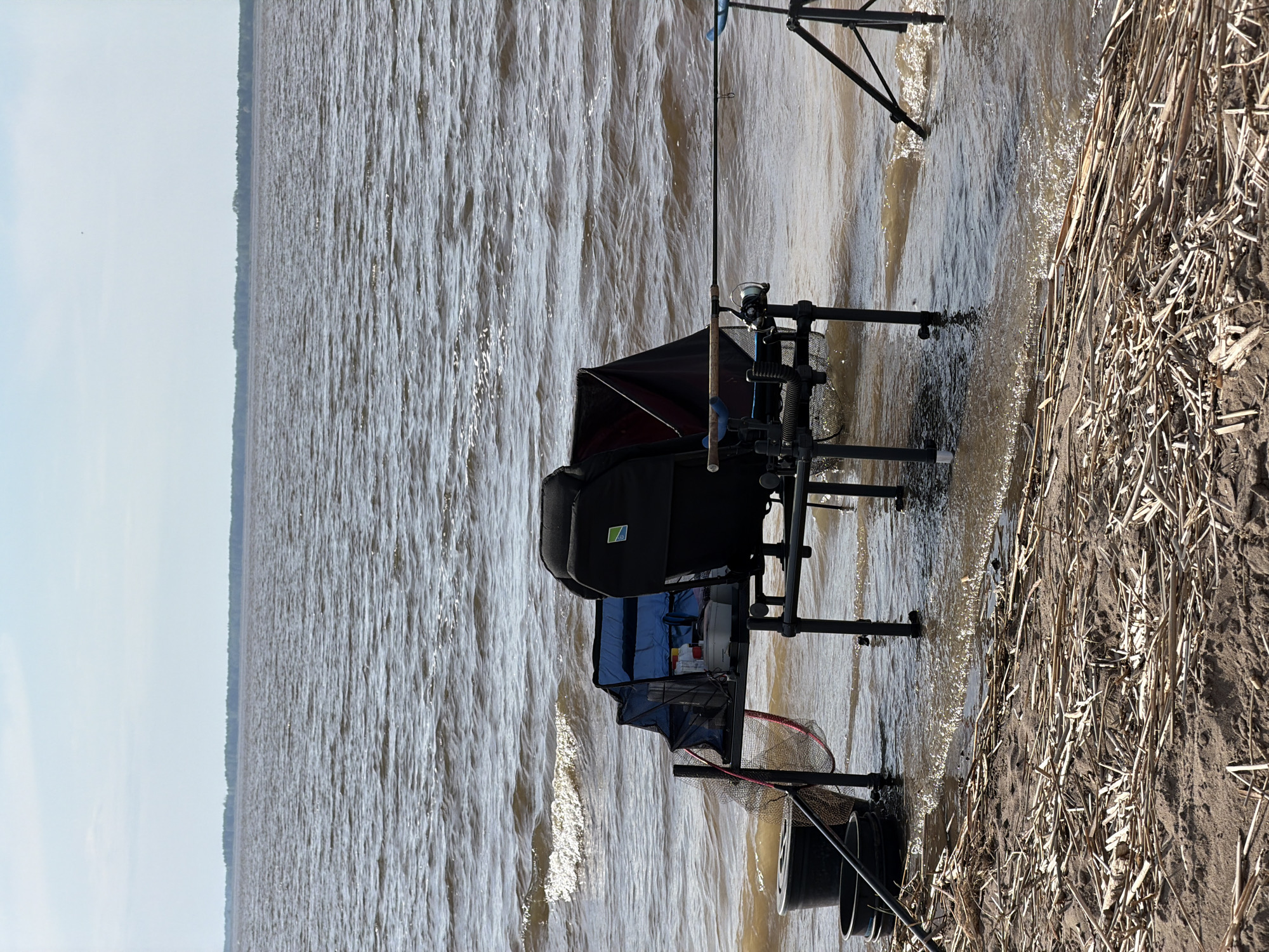 The image shows a fishing setup on a shoreline. The location is presumably in or near Krasnyi Bor, Russia. The water is choppy. A black fishing chair with attached equipment is visible, including fishing rods, a net, and buckets. The chair is positioned partly in the water, with its legs submerged. The shoreline is covered with reeds and sand. The sky is partially visible.