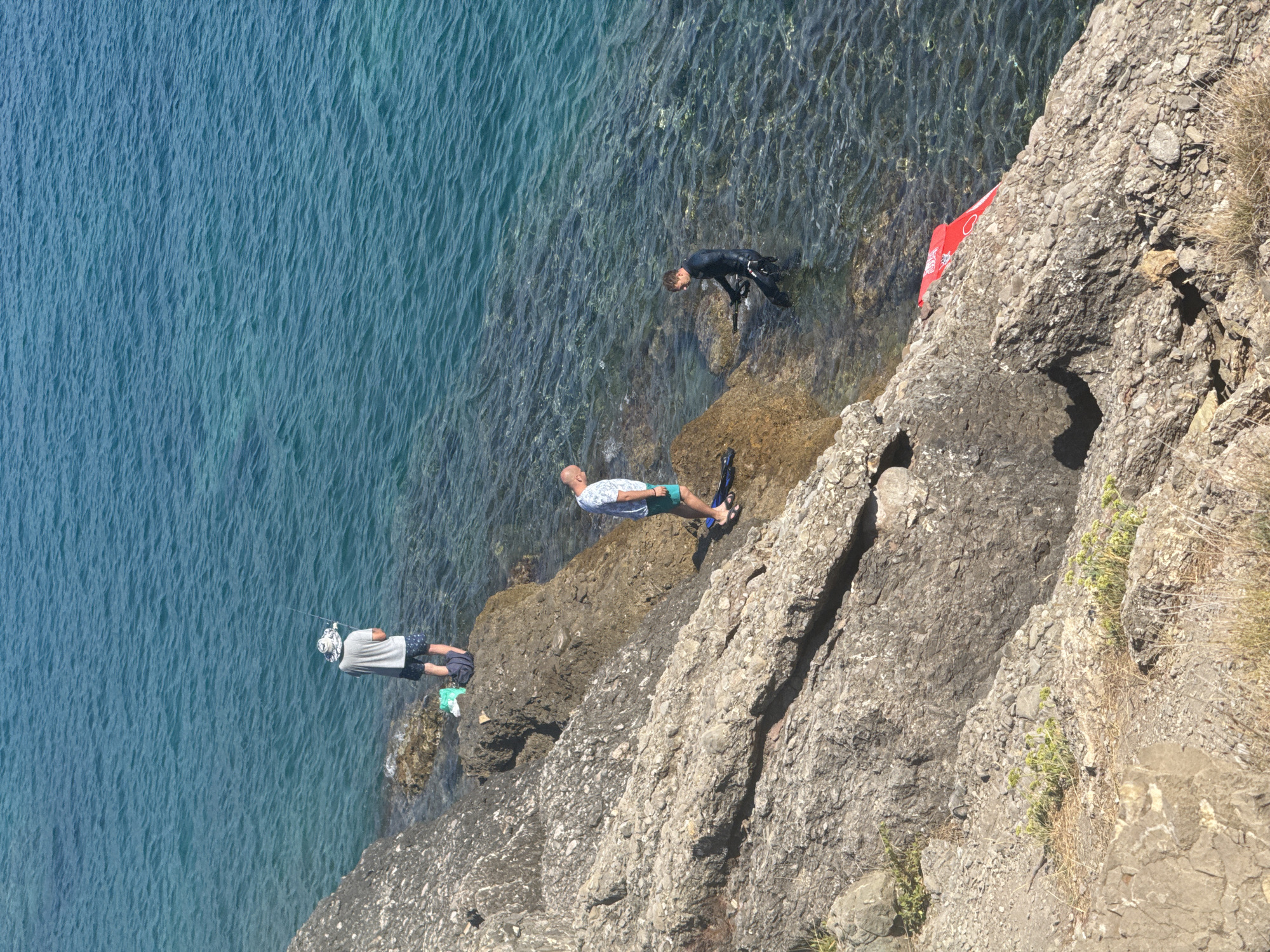 This image depicts a rocky coastline along the Adriatic Sea in Sutomore, Montenegro. In the upper left, a person wearing a hat and shorts stands on a rock, holding a fishing rod. To the right of them, a person in a blue and white shirt and shorts stands on another rock near the water's edge. Further to the right and partially in the water, a person wearing a black wetsuit, mask, and snorkel stands with a speargun. A bright red inflatable object, possibly a buoy or watercraft, is partially visible resting on the rocks near the water. The sea is a clear turquoise color, with visible ripples on the surface, and the seabed is discernible in the shallows. The terrain is characterized by rough, grey rocks and sparse vegetation.