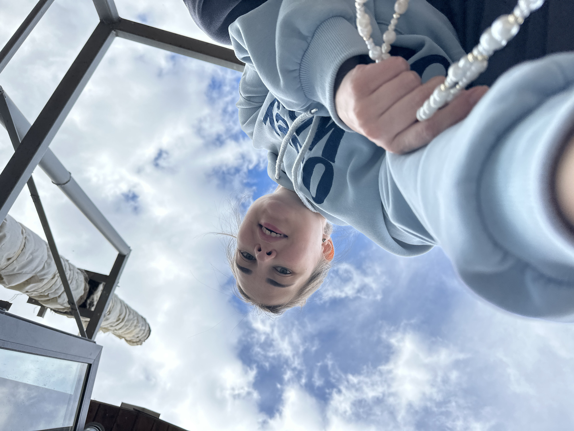 A young person wearing a light blue hooded sweatshirt with dark blue lettering and holding a string of white beads is viewed from a low angle, looking down at the camera. The background is primarily sky with scattered clouds. A metal structure is visible in the upper left corner, along with a section of a white, fabric-covered duct. The location is suggested to be in Ilyinsky, Russia, although this cannot be confirmed with only visual information.
