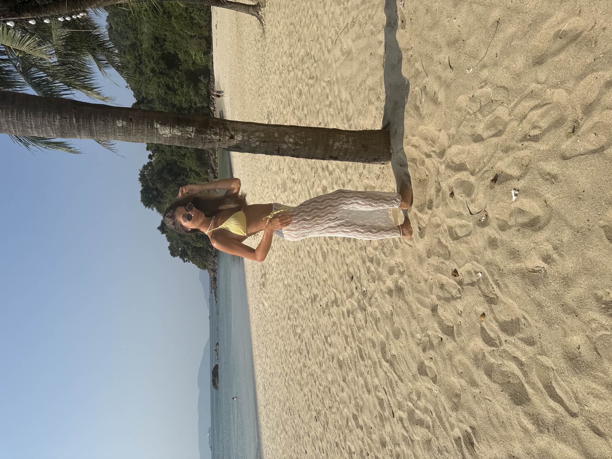A young woman stands on a sandy beach, leaning against a palm tree. She is wearing a yellow top and white pants, and sunglasses. The beach is sunny and stretches along the coastline. Palm trees and lush green vegetation are visible in the background. The ocean is calm and clear blue. The location is Đà Nẵng, Vietnam.