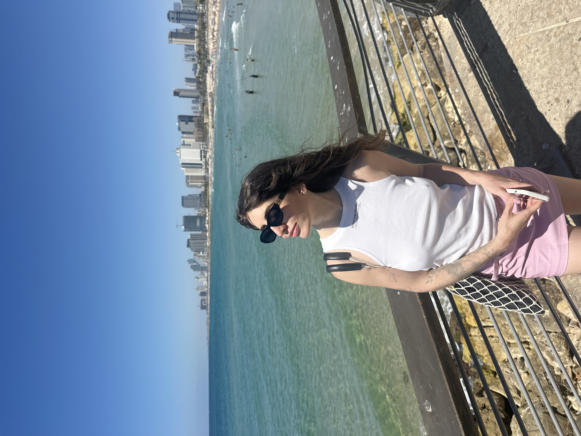 A young woman with dark hair and sunglasses is standing on a wooden promenade, looking out at the sea. She is wearing a white tank top and a pink skirt. Behind her, the Tel Aviv coastline is visible, with a clear blue sky and buildings in the distance. The water is a vibrant turquoise, with small waves and scattered beachgoers. The promenade has metal railings and rocky supports.