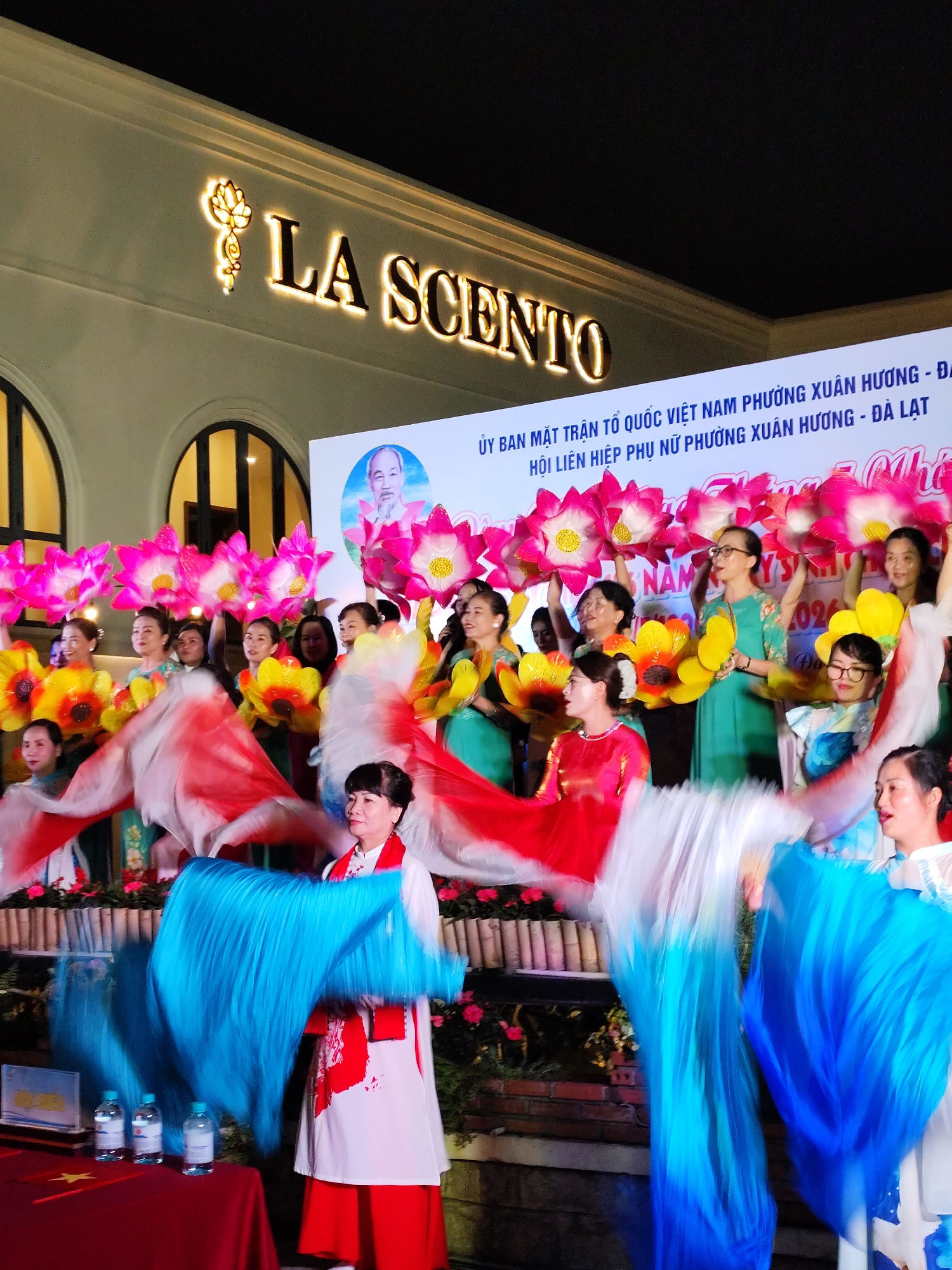 The image captures a performance in front of a building in the city of Da Lat, Vietnam, indicated by the text on a banner in the background. The building's signage reads "LA SCENTO."
A group of women is performing on what appears to be a stage. Some of the women are holding large, artificial lotus flowers in pink and yellow. In the foreground, other women are holding and swirling long, flowing silk or fabric fans in blue, red, and white colors.
The dancers are wearing traditional Vietnamese garments. A small red table covered with a red cloth sits in the left foreground, bearing a small Vietnamese flag, a plaque, and several bottles of water. A backdrop behind the performers features a portrait of Ho Chi Minh, alongside the text "ỦY BAN MẶT TRẬN TỔ QUỐC VIỆT NAM PHƯỜNG XUÂN HƯƠNG - ĐI" and "HỘI LIÊN HIỆP PHỤ NỮ PHƯỜNG XUÂN HƯƠNG - ĐÀ LẠT."
