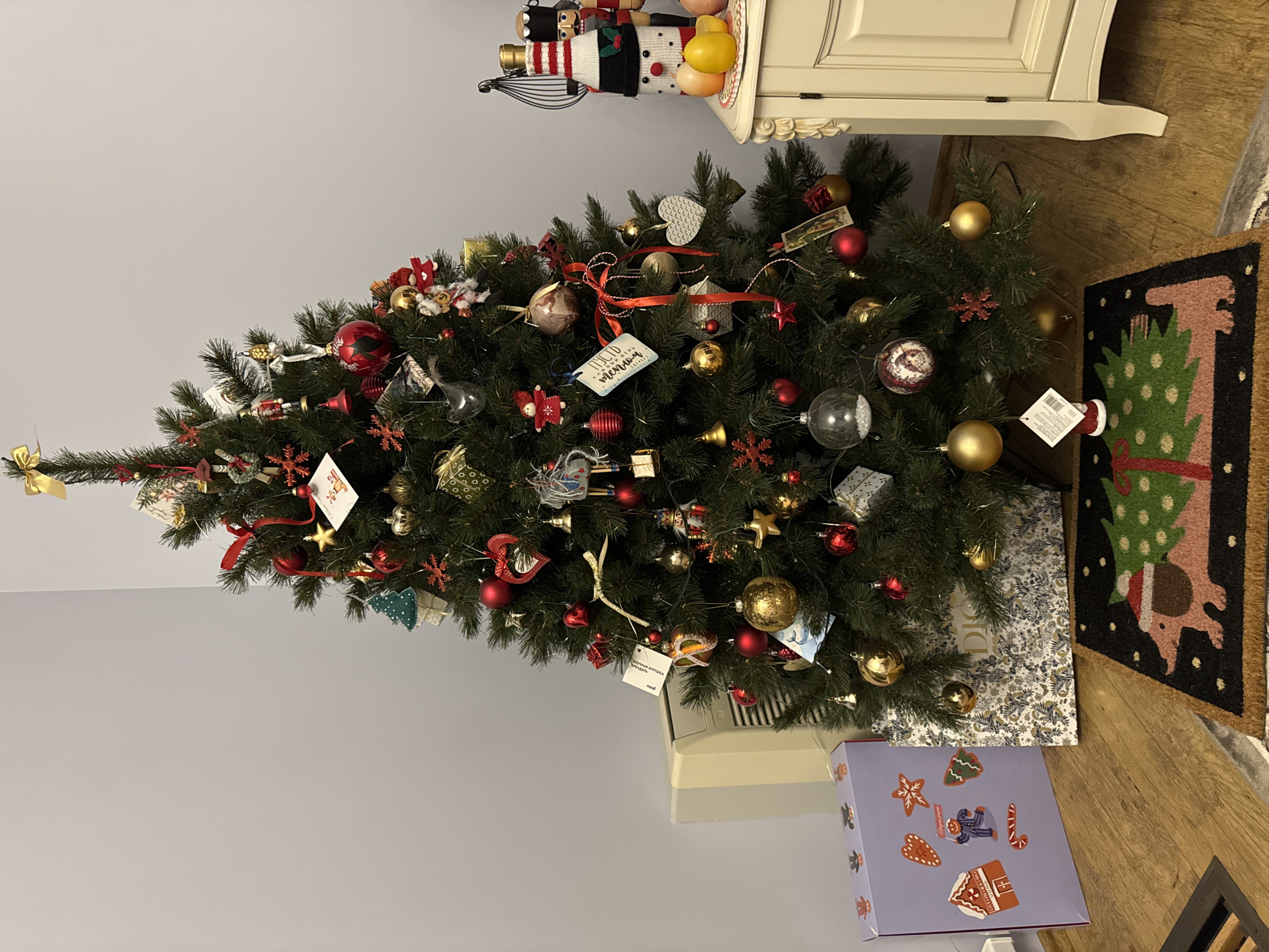 An indoor Christmas tree is decorated with red and gold ornaments, small figures, and lights. A gold ribbon bow tops the tree. To the left of the tree, a beige air conditioner is visible, and a lavender gift box with gingerbread-themed designs sits on a wooden floor. A patterned gift bag is placed beneath the tree. To the right of the tree, a white cabinet displays a snowman-decorated bottle, two fruit, and a nutcracker figure. A doormat with a dachshund carrying a Christmas tree is in front of the tree. The scene appears to be in a residential setting in Yeysk, Russia.