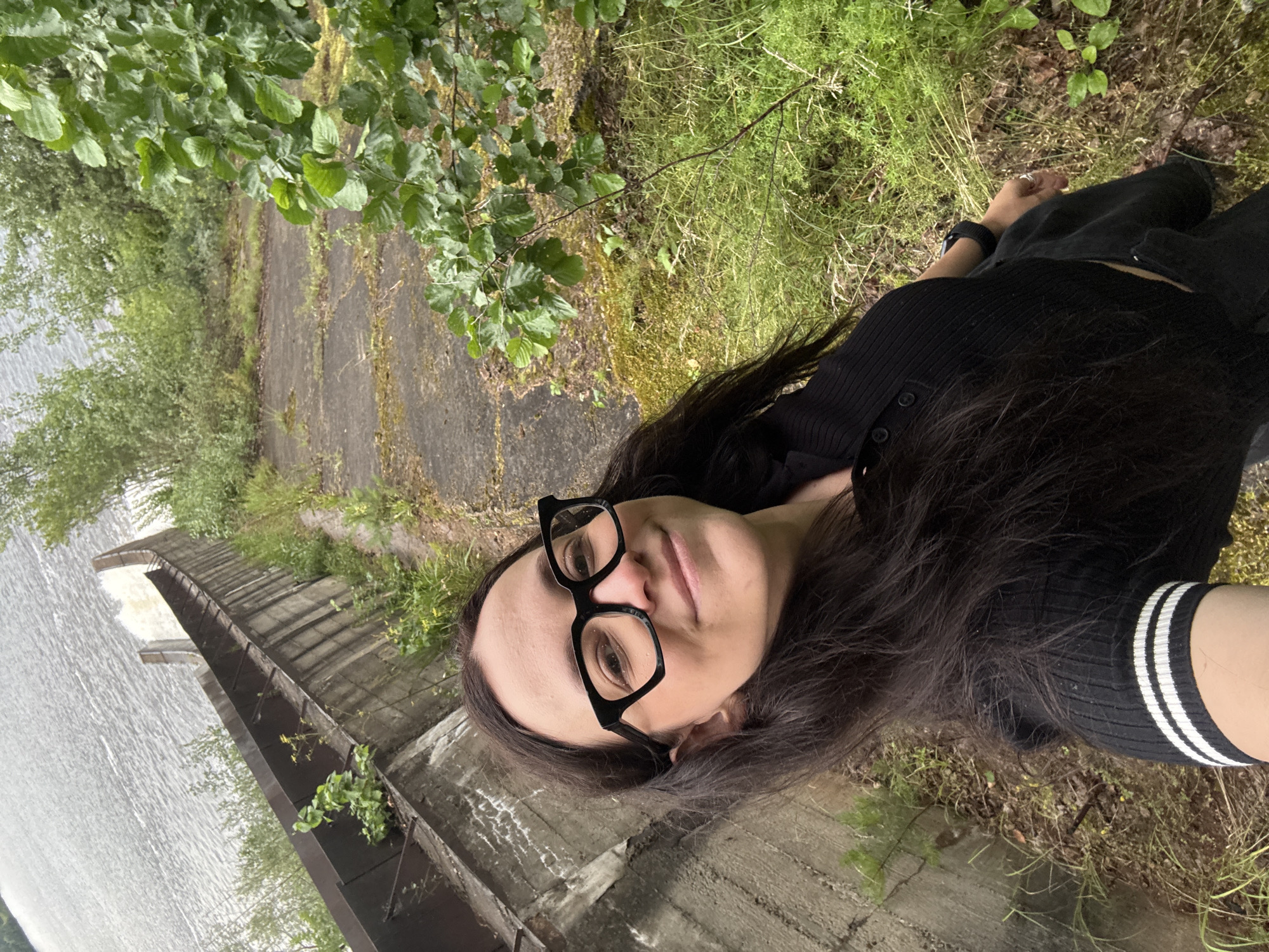 A woman wearing black-rimmed glasses and a black collared shirt with white stripes on the sleeves stands by a concrete structure and a body of water. The structure appears to be part of a dam or spillway, with water flowing over its edge in the background. The surrounding environment is overgrown with green vegetation, including trees and grass. The scene is captured from a high angle, looking down towards the woman. The location is identified as Chernushka, Russia.