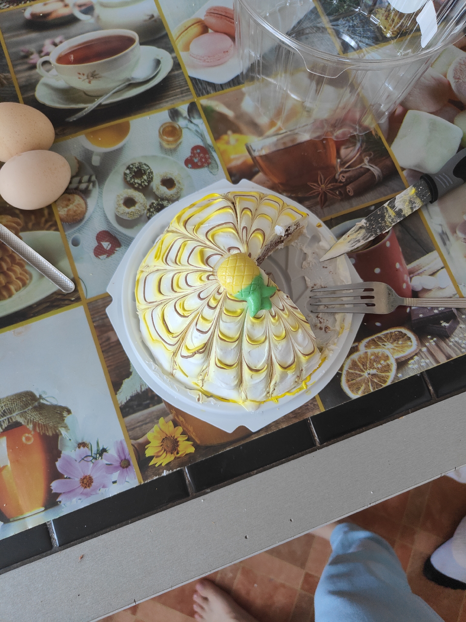 The image shows a partially eaten cake on a table. The cake is decorated with white and yellow icing in a swirling pattern, and has a pineapple-shaped decoration on top. A fork and a knife are placed next to the cake, suggesting that it has been eaten. The table surface has a printed design that features images of tea cups, macarons, donuts, honey jars, and flowers. There are two eggs in the upper left corner of the image. A clear plastic container is visible in the upper right corner. The image is taken from a top-down perspective. The table appears to be set on a patterned floor, and a person's leg and foot are visible in the lower right portion of the image, wearing blue pants and a white sock. There are no identifiable landmarks or environmental features that would indicate a specific location in Zheleznogorsk, Russia.