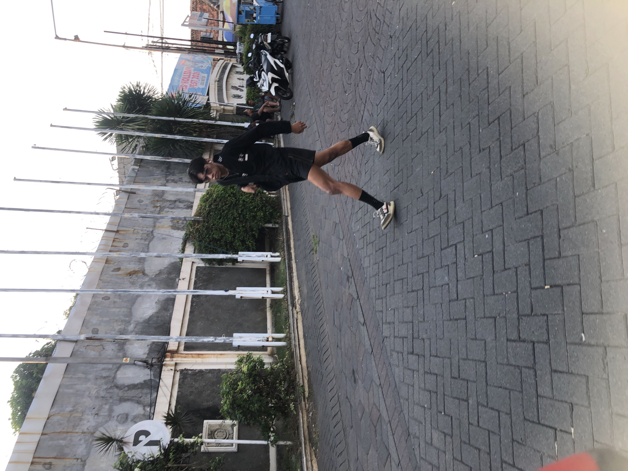 A young man wearing sunglasses, a black jacket, black shorts, black socks, and athletic shoes is walking on a paved street. He has dark hair and appears to be wearing earbuds. The street is paved with dark grey interlocking bricks laid in a herringbone pattern. In the background, there is a concrete wall with a parking sign on the left. Several tall, thin poles stand in front of the wall. Beyond the poles, there are trees, a building with advertisements, parked motorcycles, and a blue and yellow cart. The scene appears to be a street in Boro, Indonesia, likely during the day given the lighting.